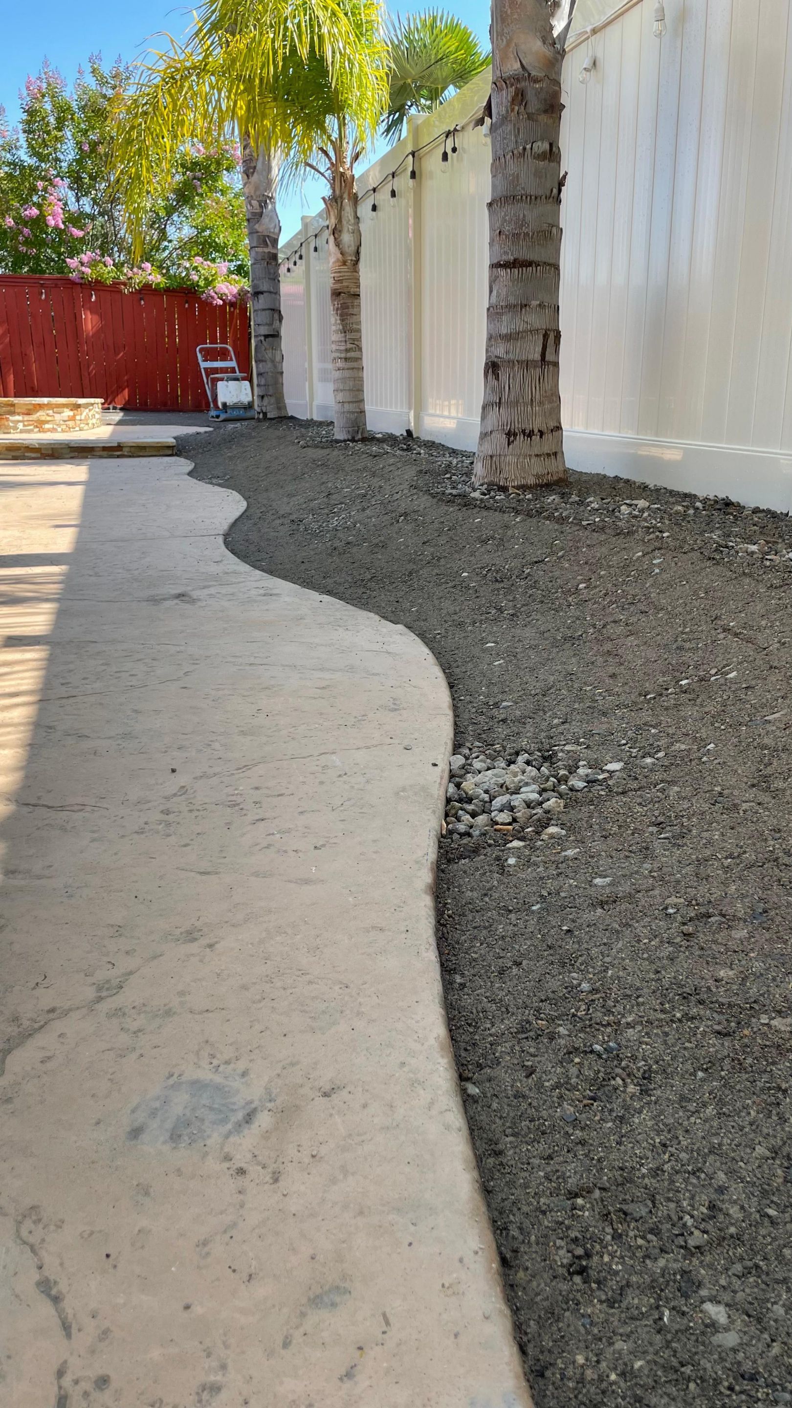 Concrete walkway curves to gravel bed with trees along a white wall.