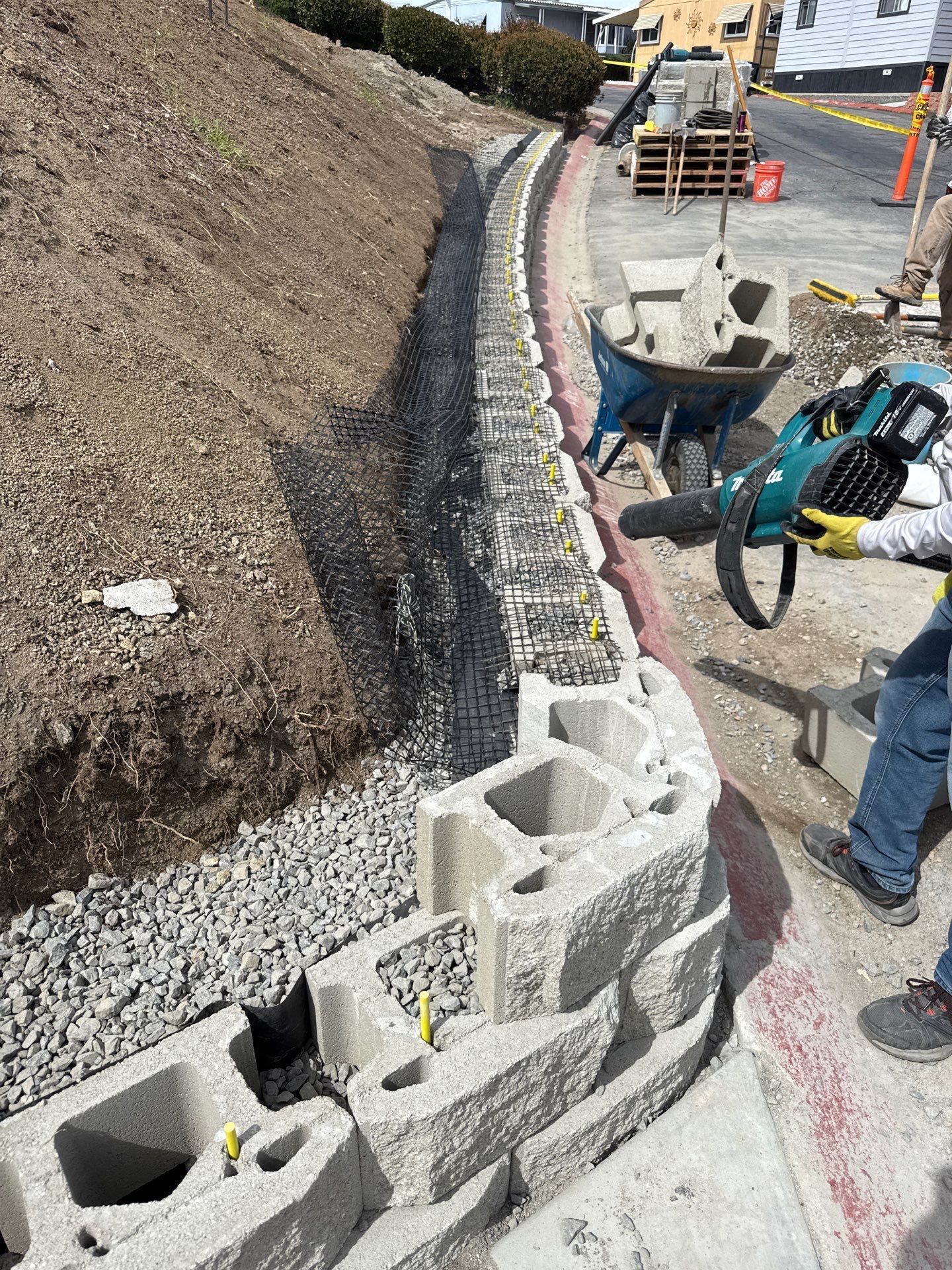 Construction of a retaining wall. Worker uses a blower. Blocks, gravel, and hillside are visible.