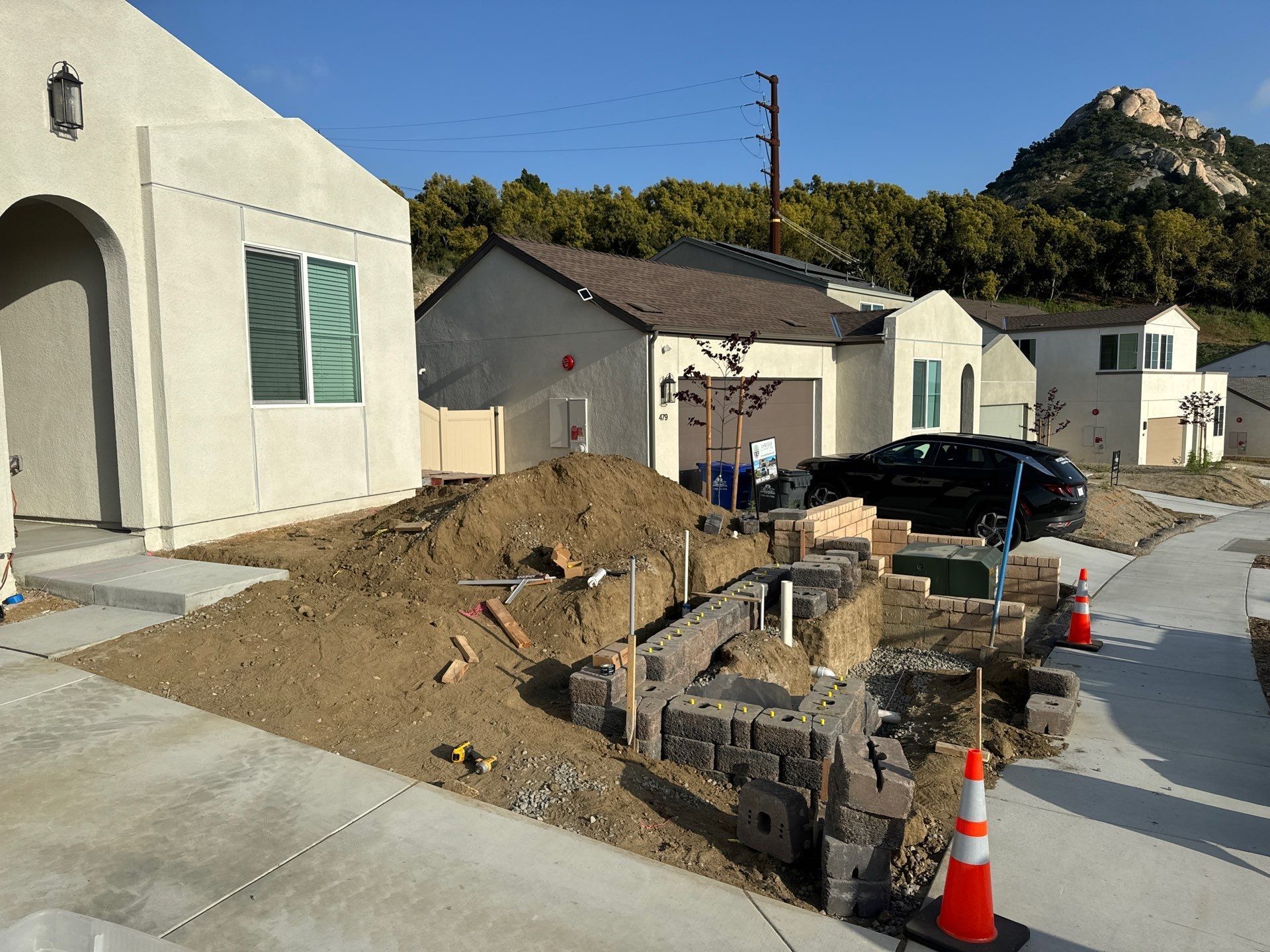 Construction site in front of houses with retaining wall being built. Dirt and tools present, mountains in background.