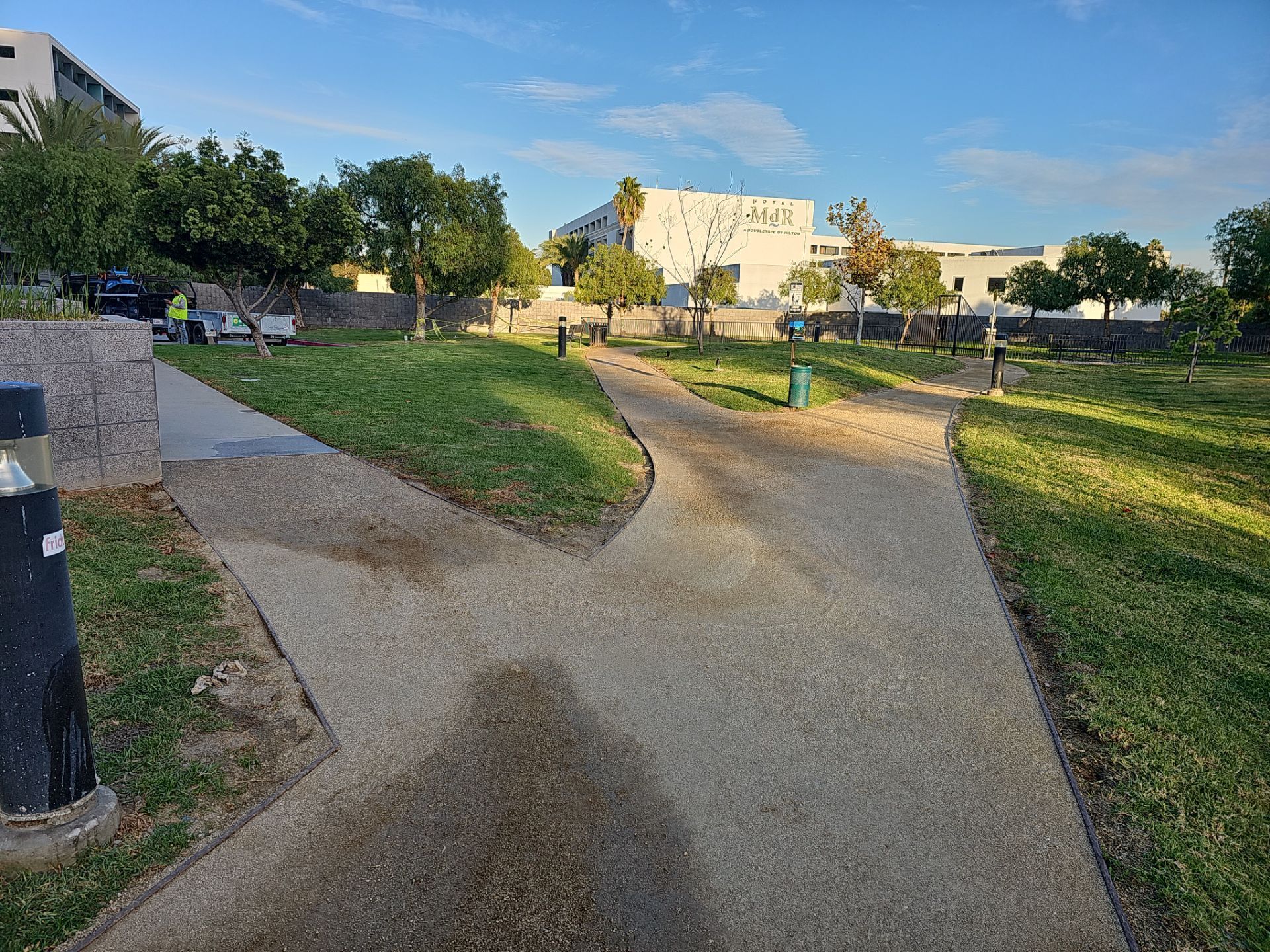 Forking paved paths in a park with green grass, trees, and a white building under a blue sky.