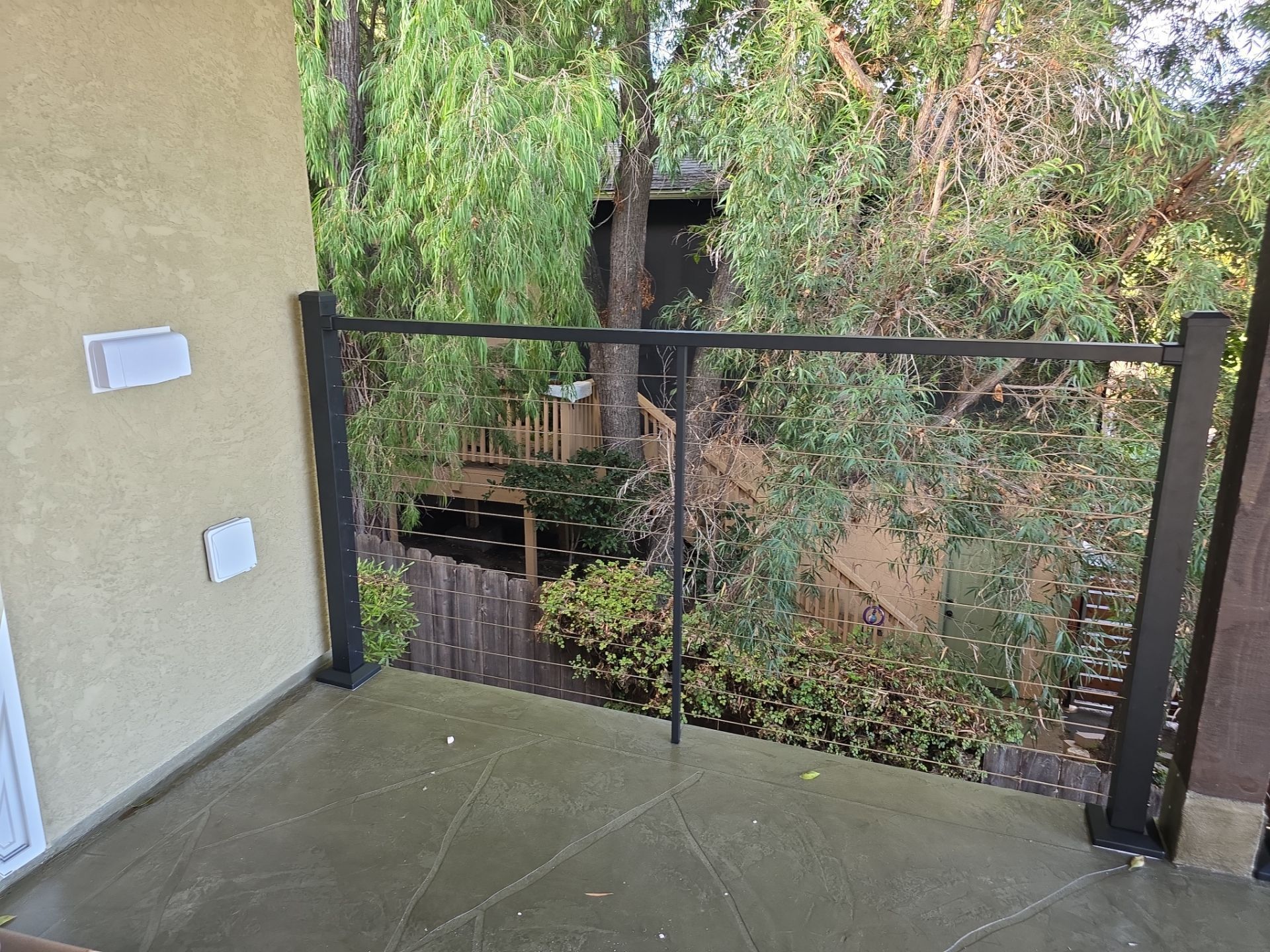Balcony with black railing, overlooking trees and a deck. Concrete floor, stucco wall.