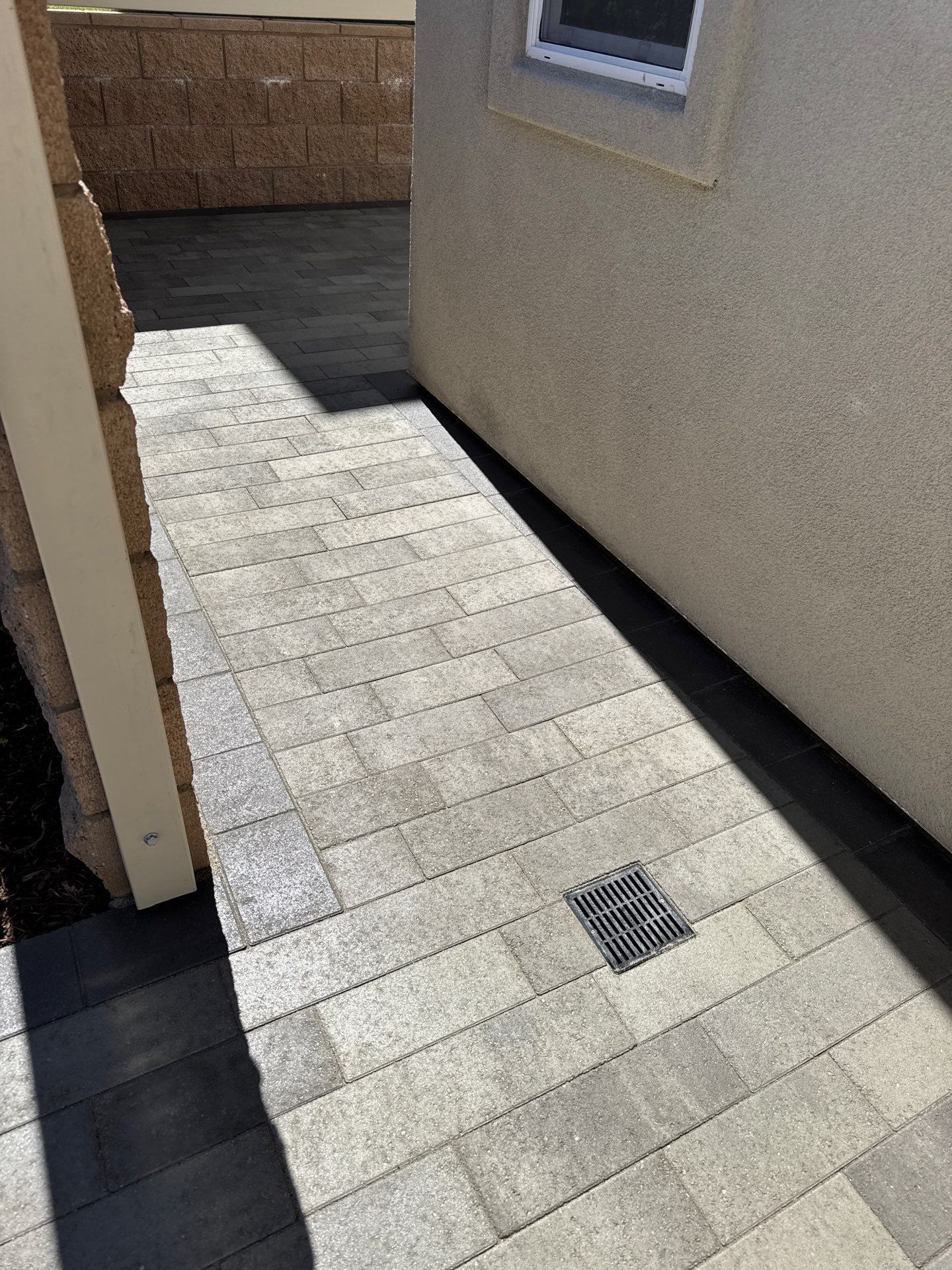 Paved walkway next to a building, with a drain, leading towards a wall.