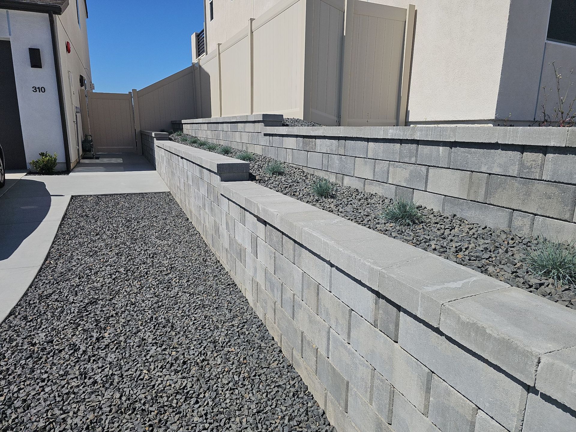Stone retaining walls with gravel and sparse plants alongside a walkway leading to a building.