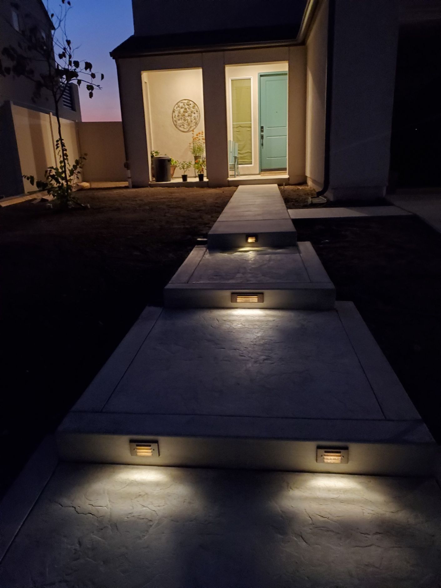 Lit concrete walkway leading to a house entrance at dusk. Embedded lights illuminate the path.