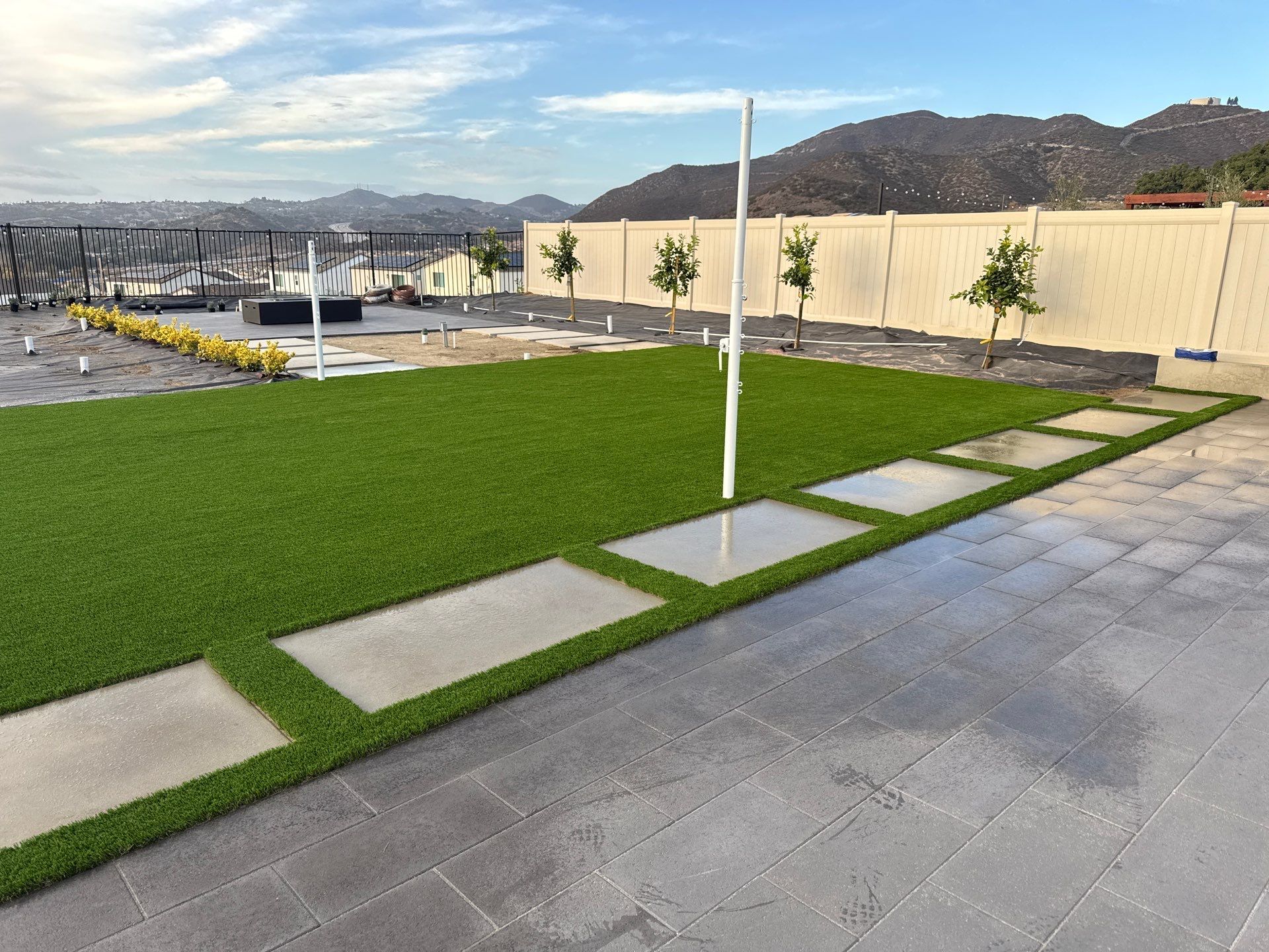 Backyard with green turf, stepping stones, gray patio, and beige fence; mountains in background.