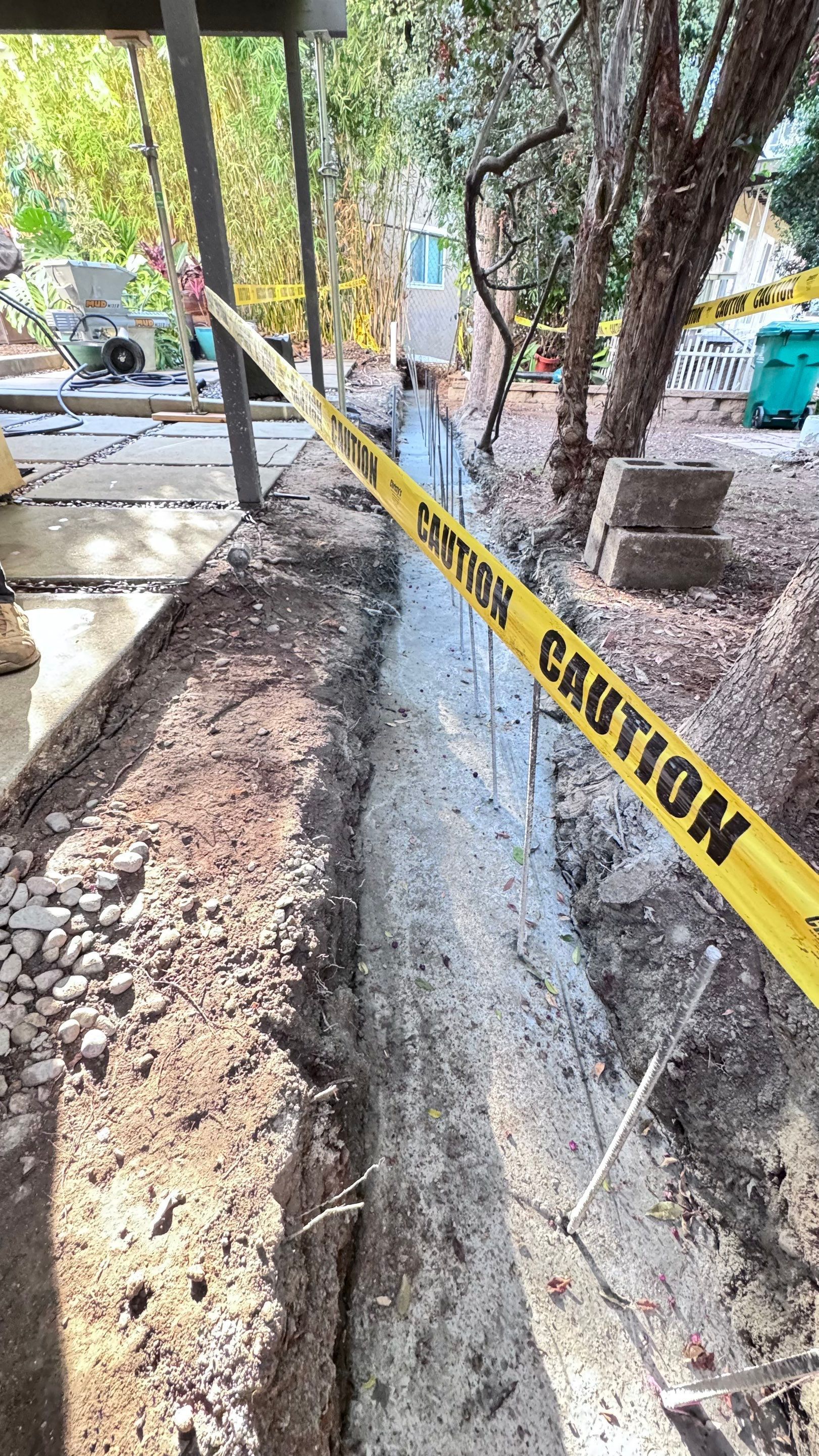 Trench dug in dirt, filled with concrete, yellow CAUTION tape across. Trees and building visible.