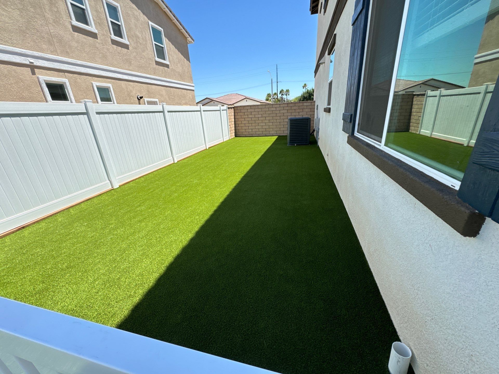 Green artificial turf backyard between a white fence and a beige house, under a blue sky.