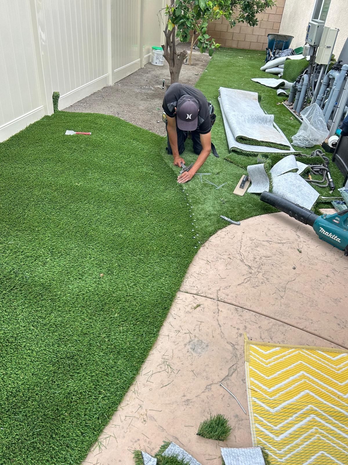 Person installing artificial turf in a backyard. Green turf, gray concrete, white fence.