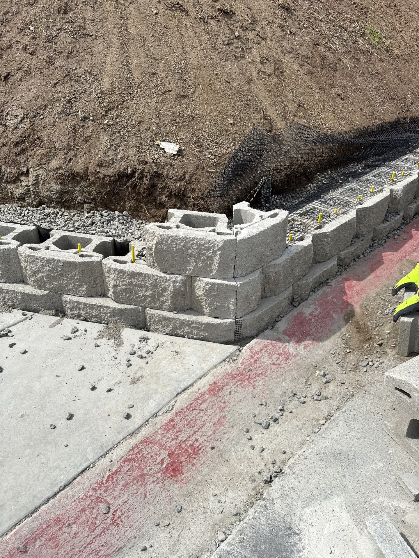 Retaining wall under construction with gray concrete blocks. Soil bank in background. Concrete sidewalk in foreground.