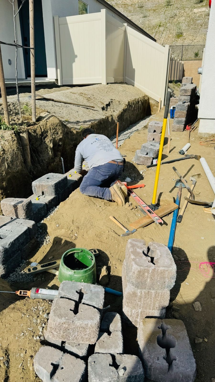 Man installing paving stones in a trench near a fence. Earth tones dominate. Tools and materials are scattered.