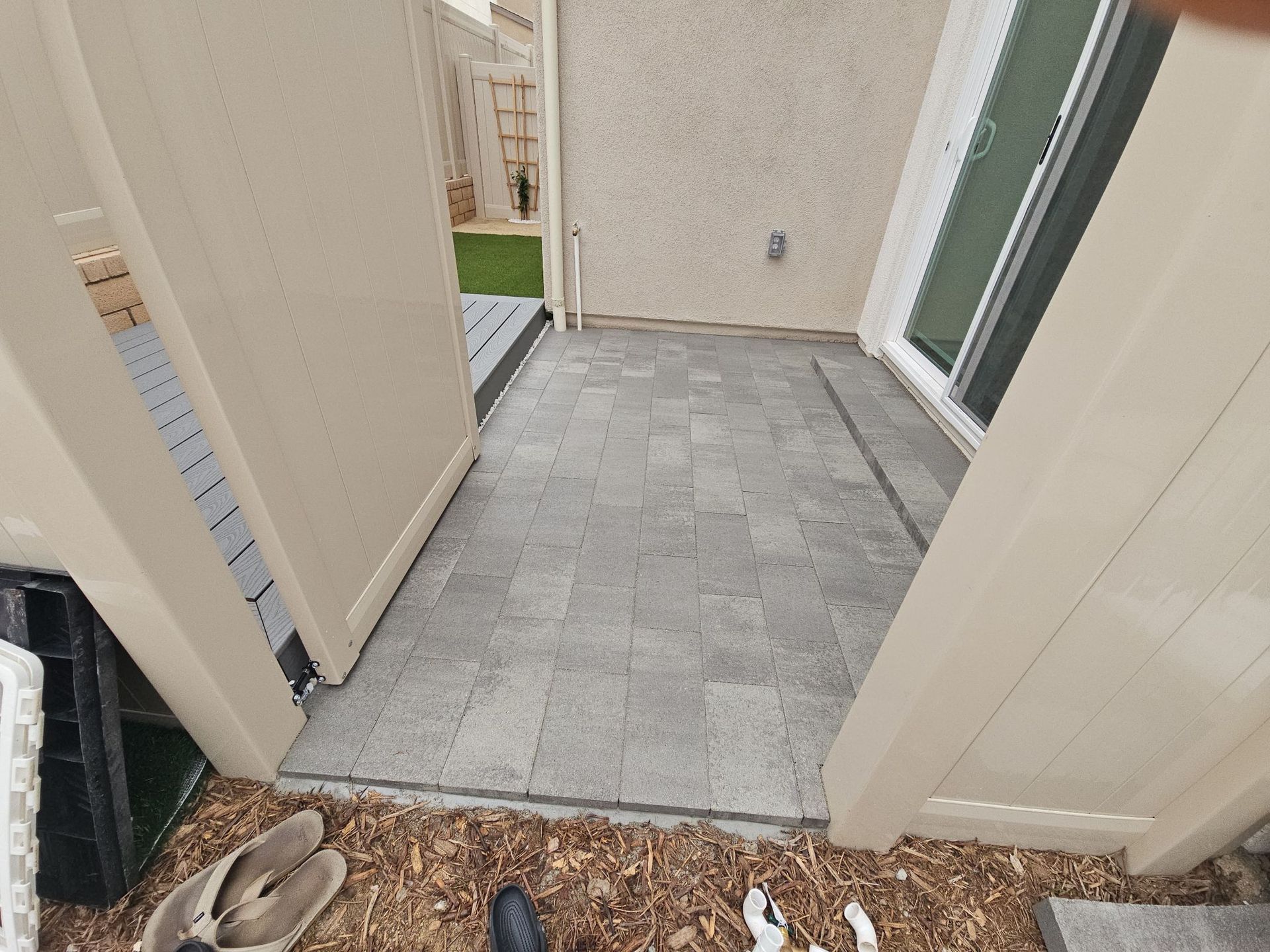 A patio paved with gray bricks, between two beige fence sections. A sliding glass door is on the right.