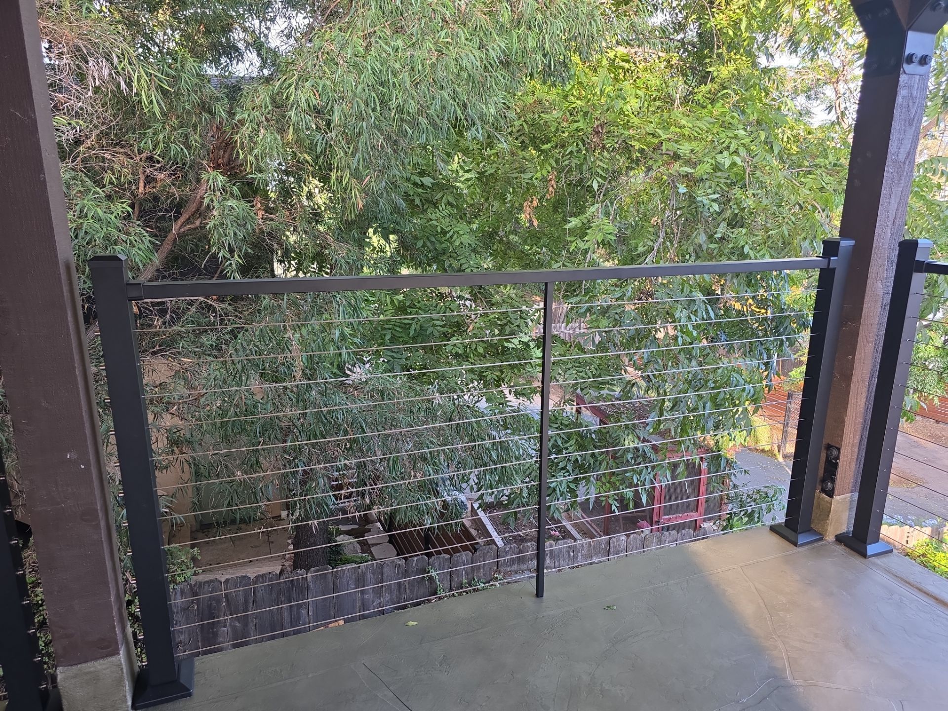 Balcony with black cable railing and trees in the background. Brown posts, green foliage.