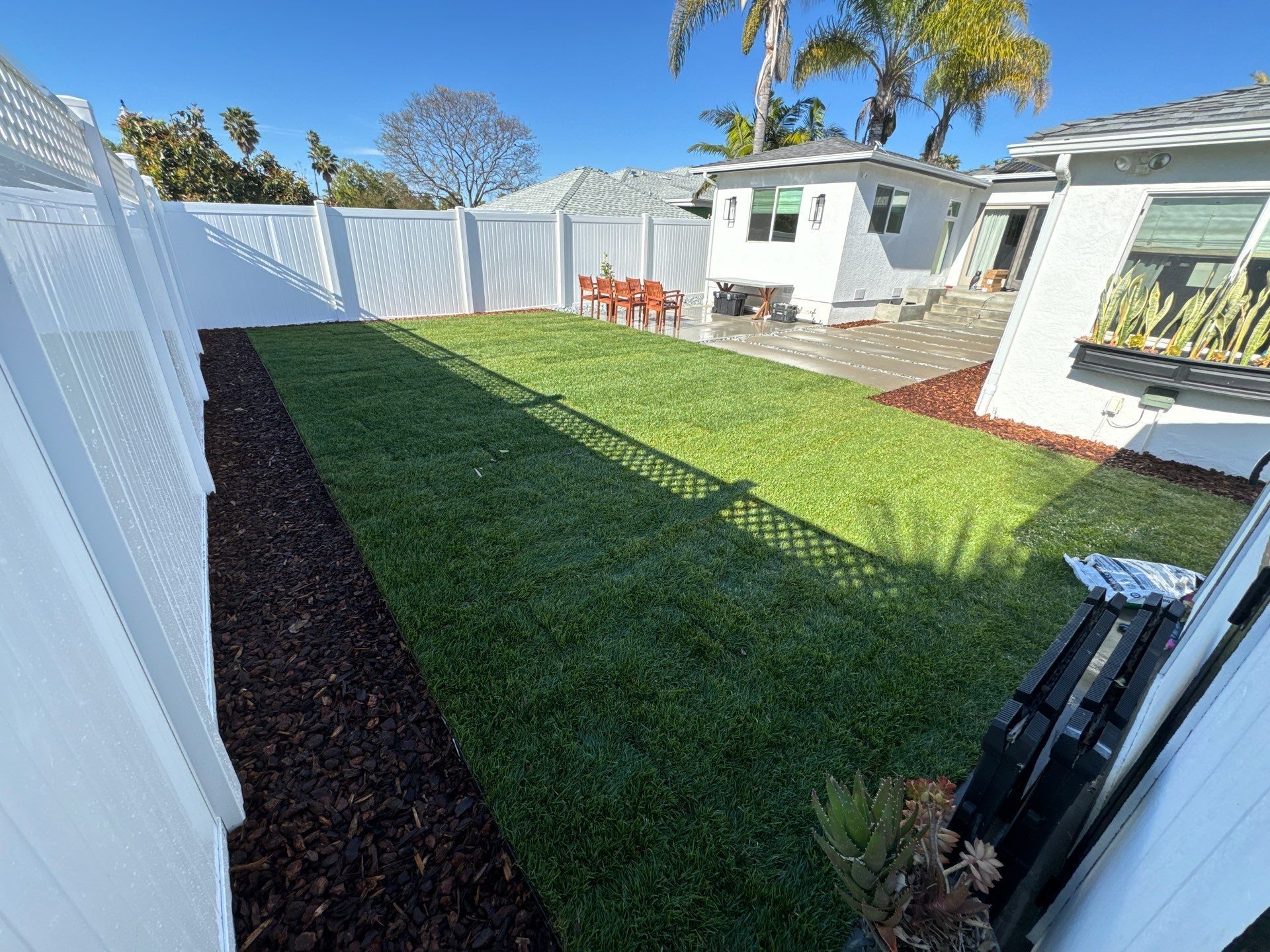 Lawn with white fence, brown mulch border, and small shed, under a bright blue sky.