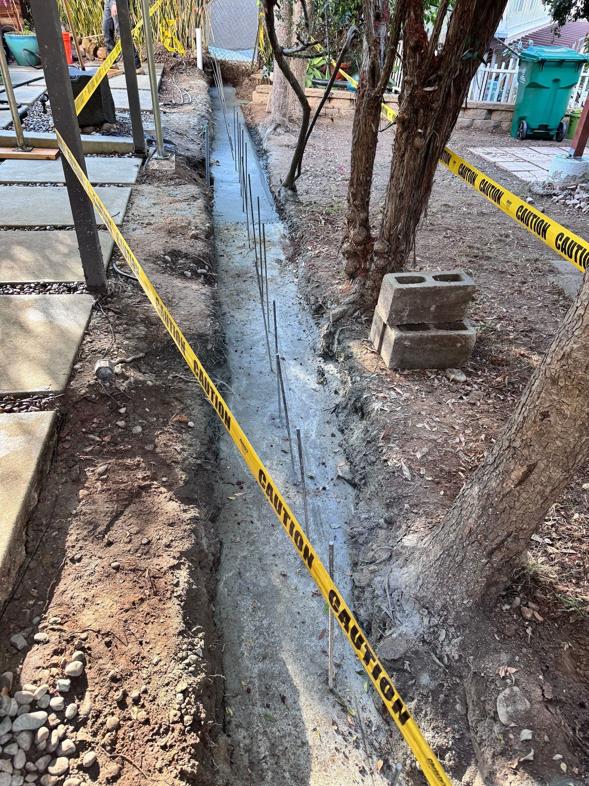 A narrow trench with wet concrete and rebar, marked with yellow caution tape, alongside a stone walkway.