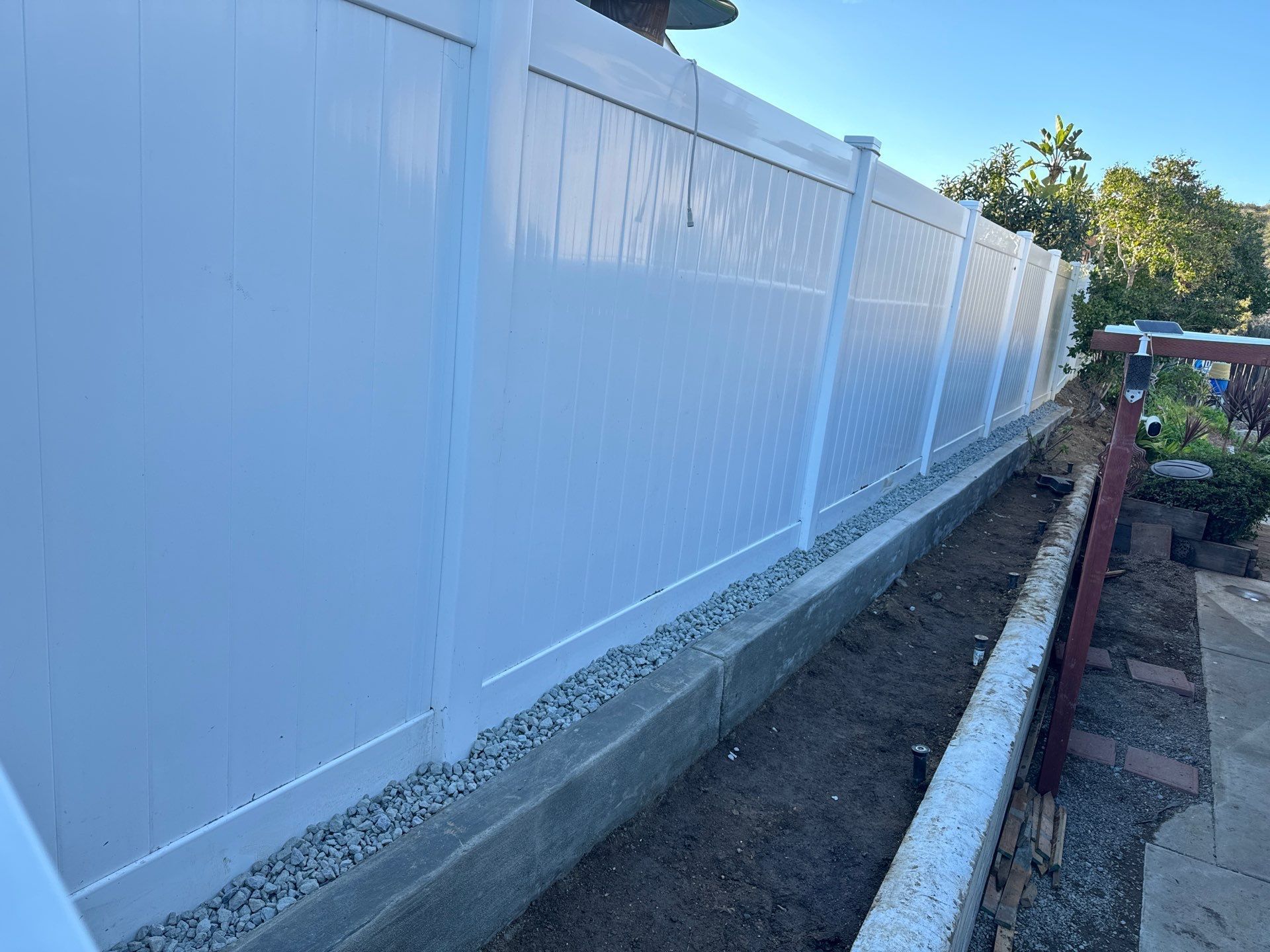 White vinyl fence next to a concrete and gravel border on a sunny day.