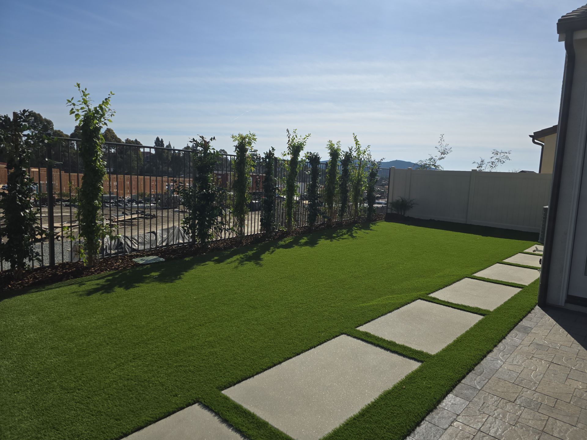 Green lawn with stepping stones, a line of tall green plants, and a fence under a blue sky.