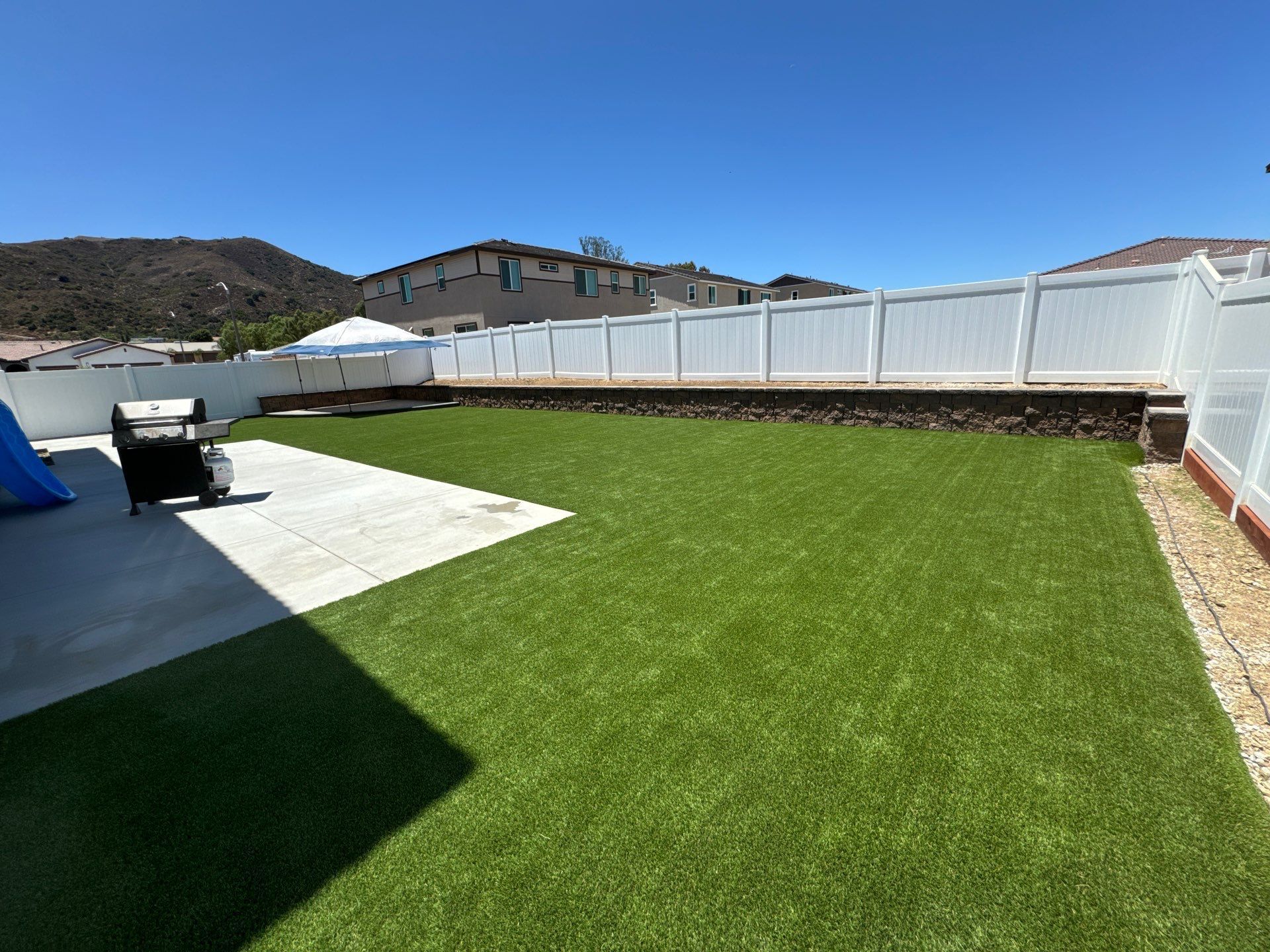 Backyard with green turf, white fence, concrete patio, and a grill on a sunny day.