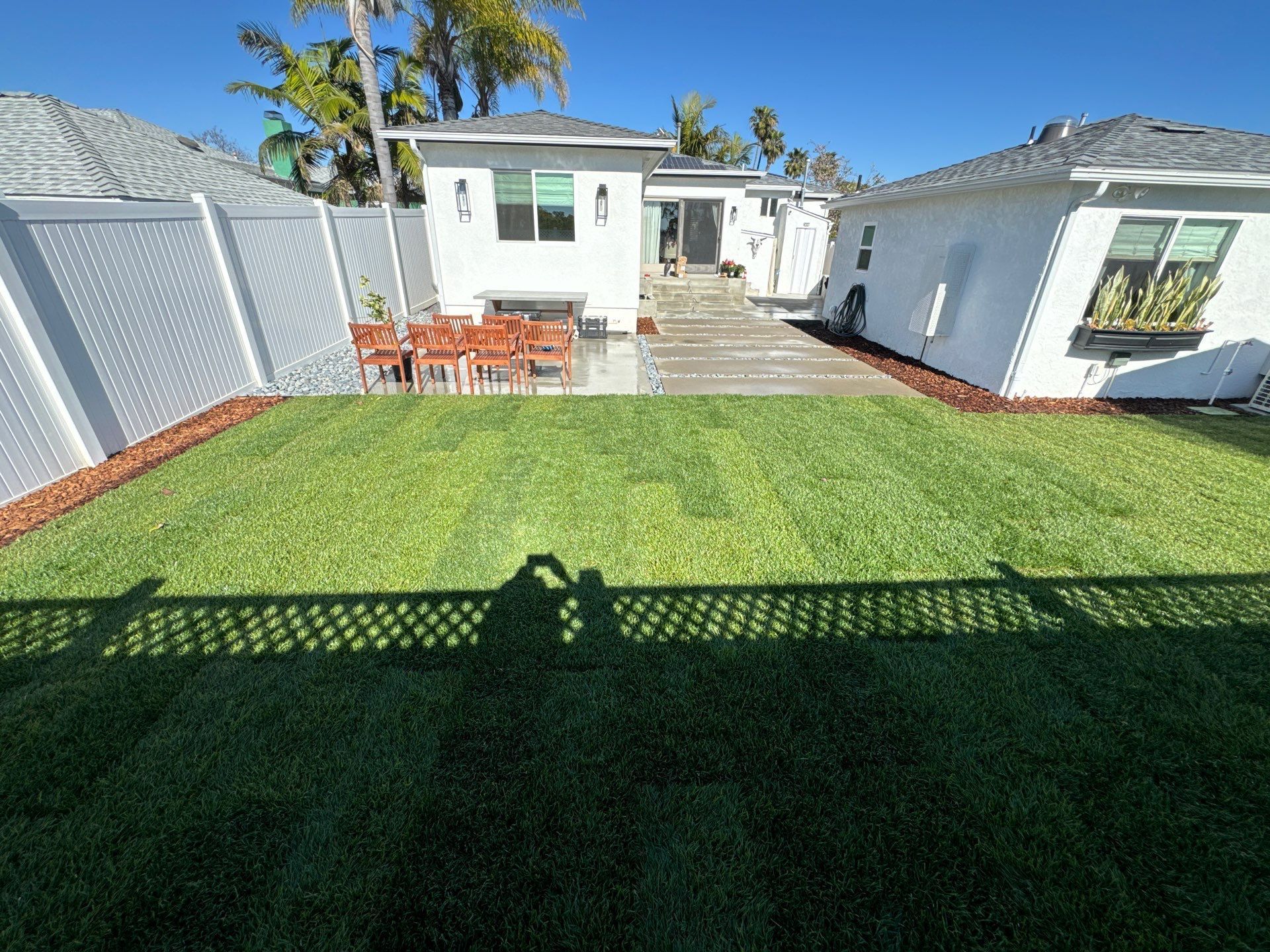 Backyard with green lawn, white fence, patio, and two houses under a blue sky.
