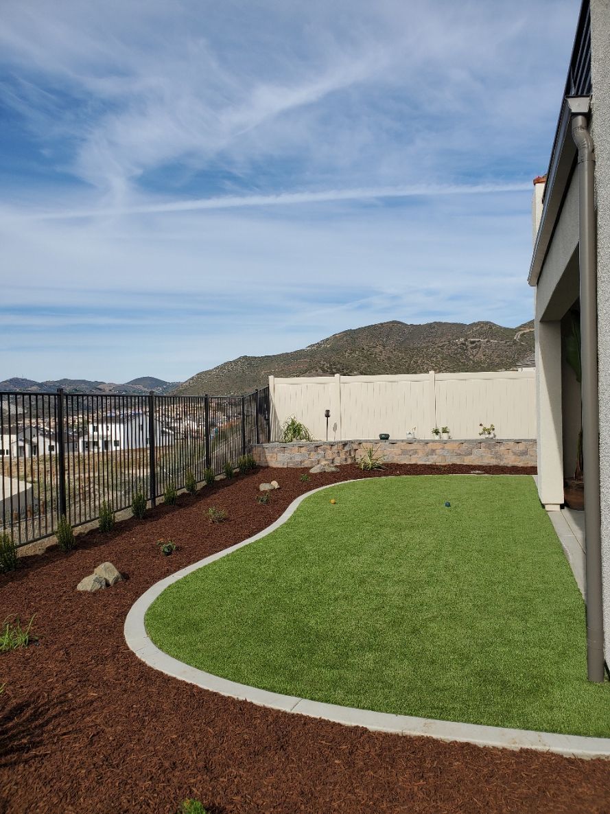 Backyard with green lawn, mulch border, fence, and hills in the distance under a blue sky.