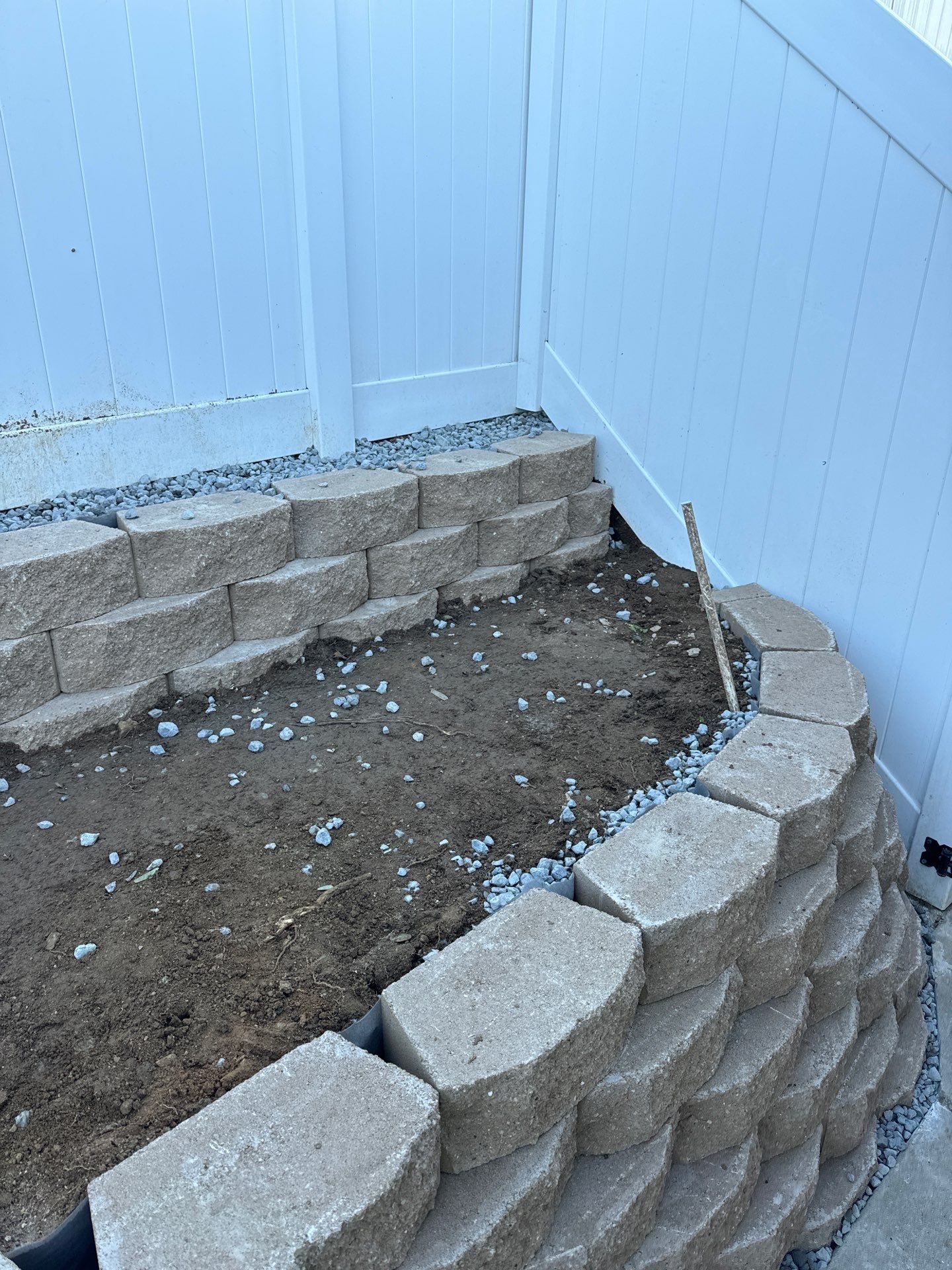 Retaining wall constructed with stacked, gray concrete blocks, beside a white vinyl fence. Garden bed area.
