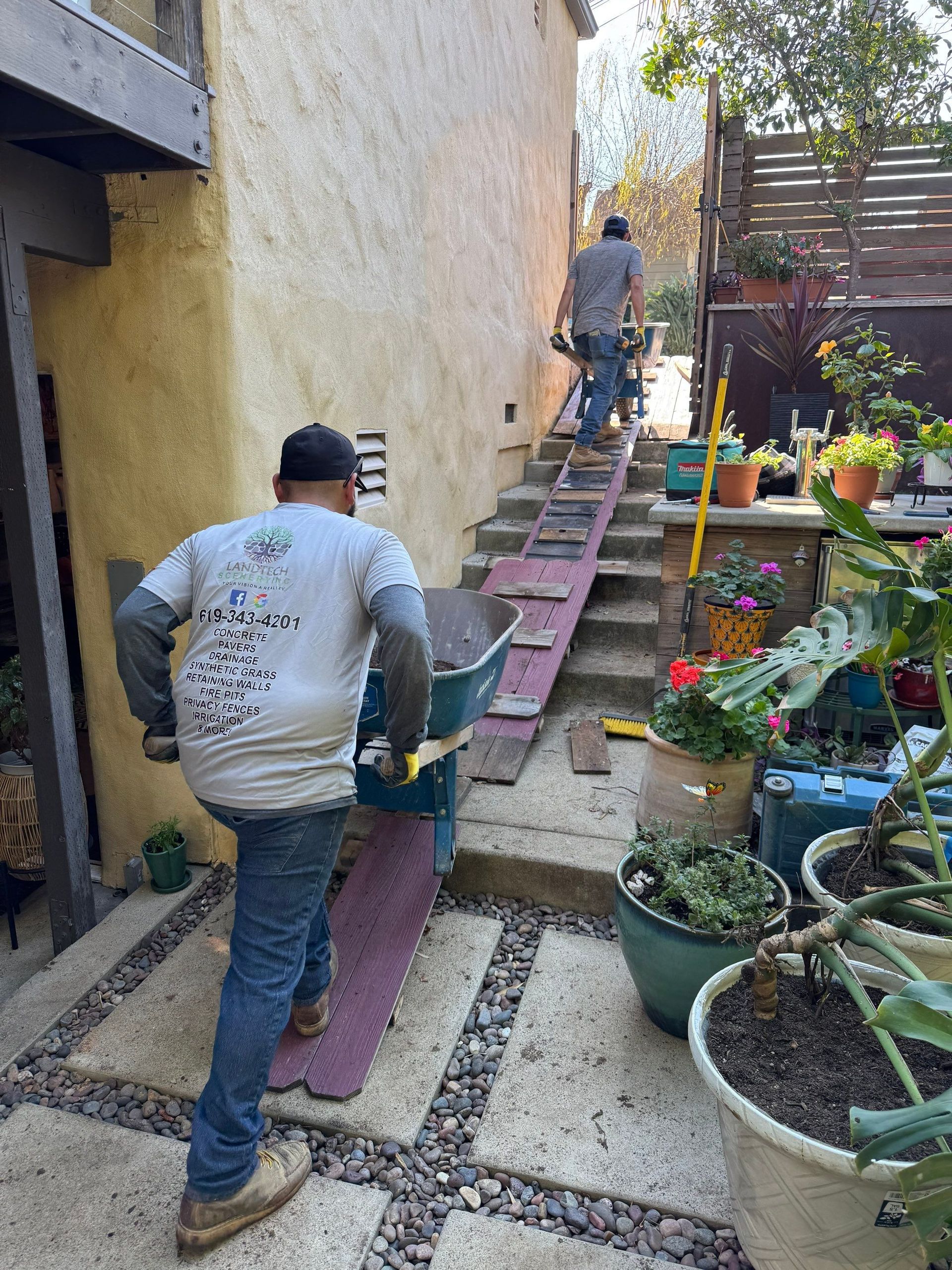 Two men carrying equipment up stairs using ramps, one with a wheelbarrow. Outdoor setting.