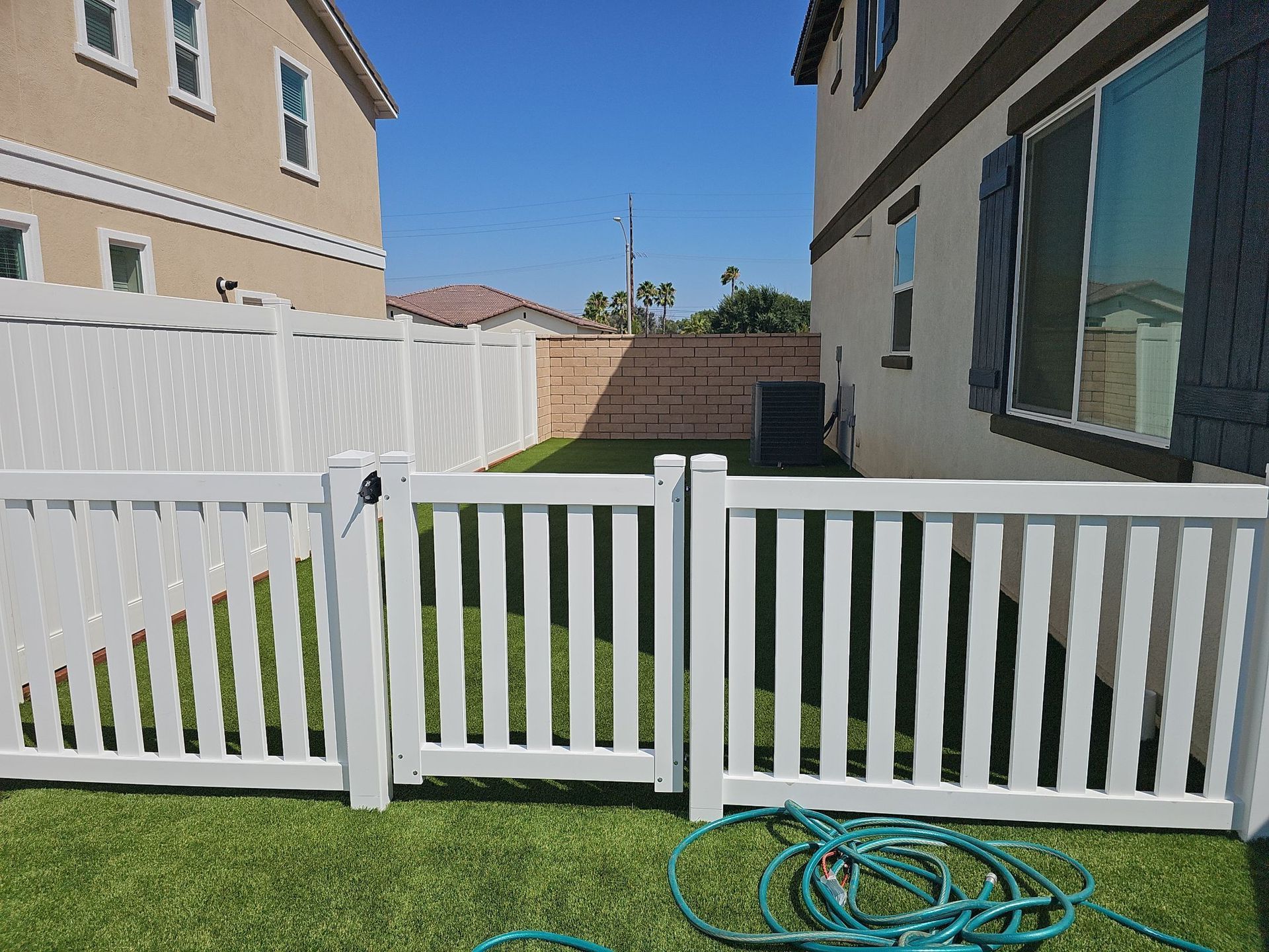 White picket fence with gate in front of a backyard with grass, houses, and blue sky.