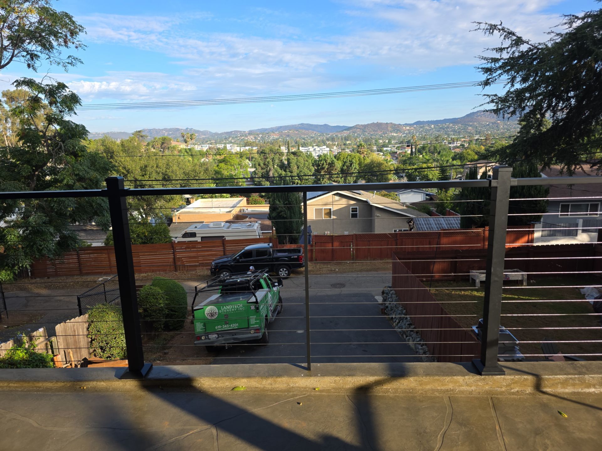 View overlooking a parking area with vehicles, houses, and trees against a backdrop of distant hills and a blue sky.