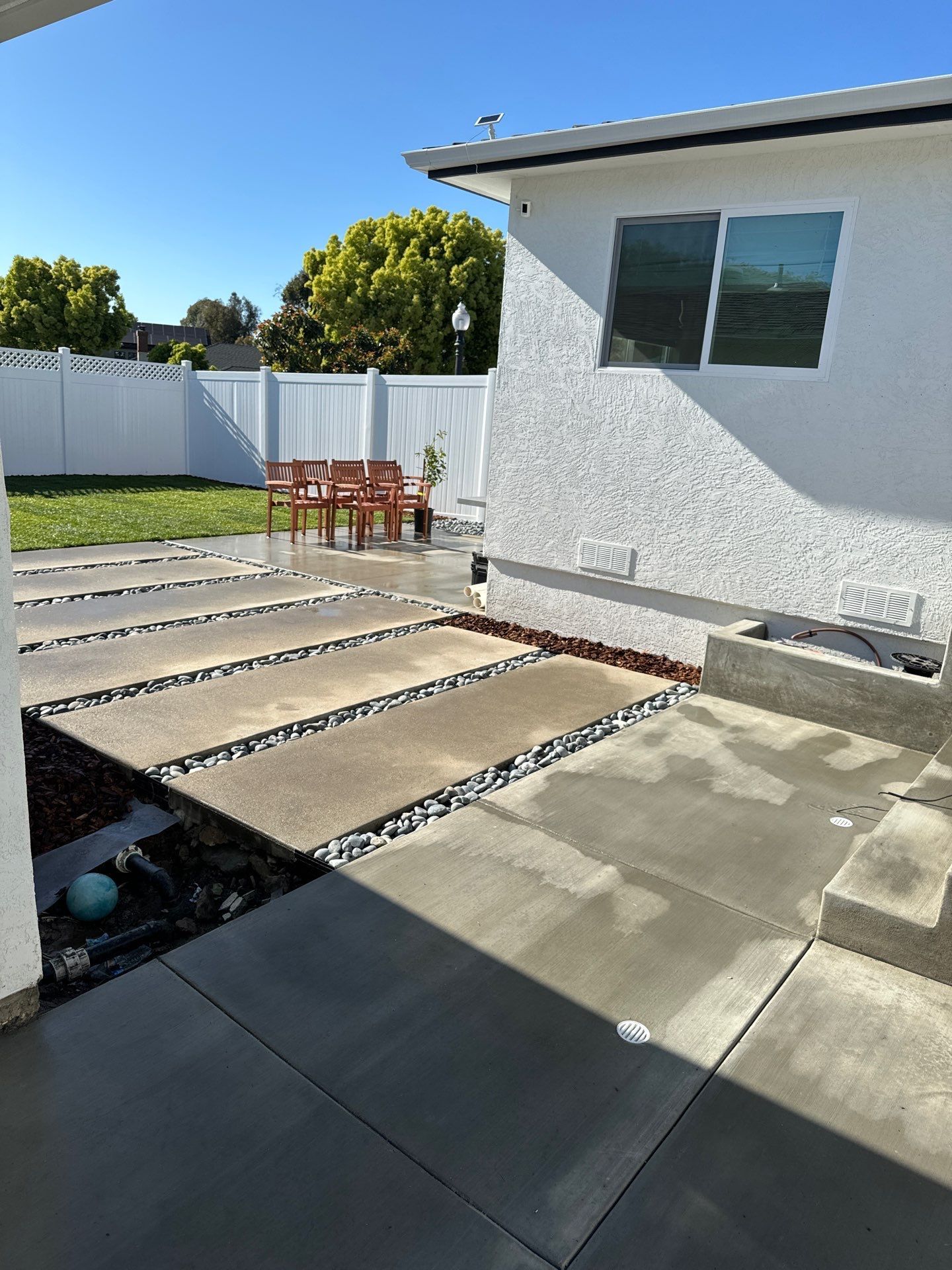 Concrete patio with stone path leading to a seating area in a backyard. White fence and house.