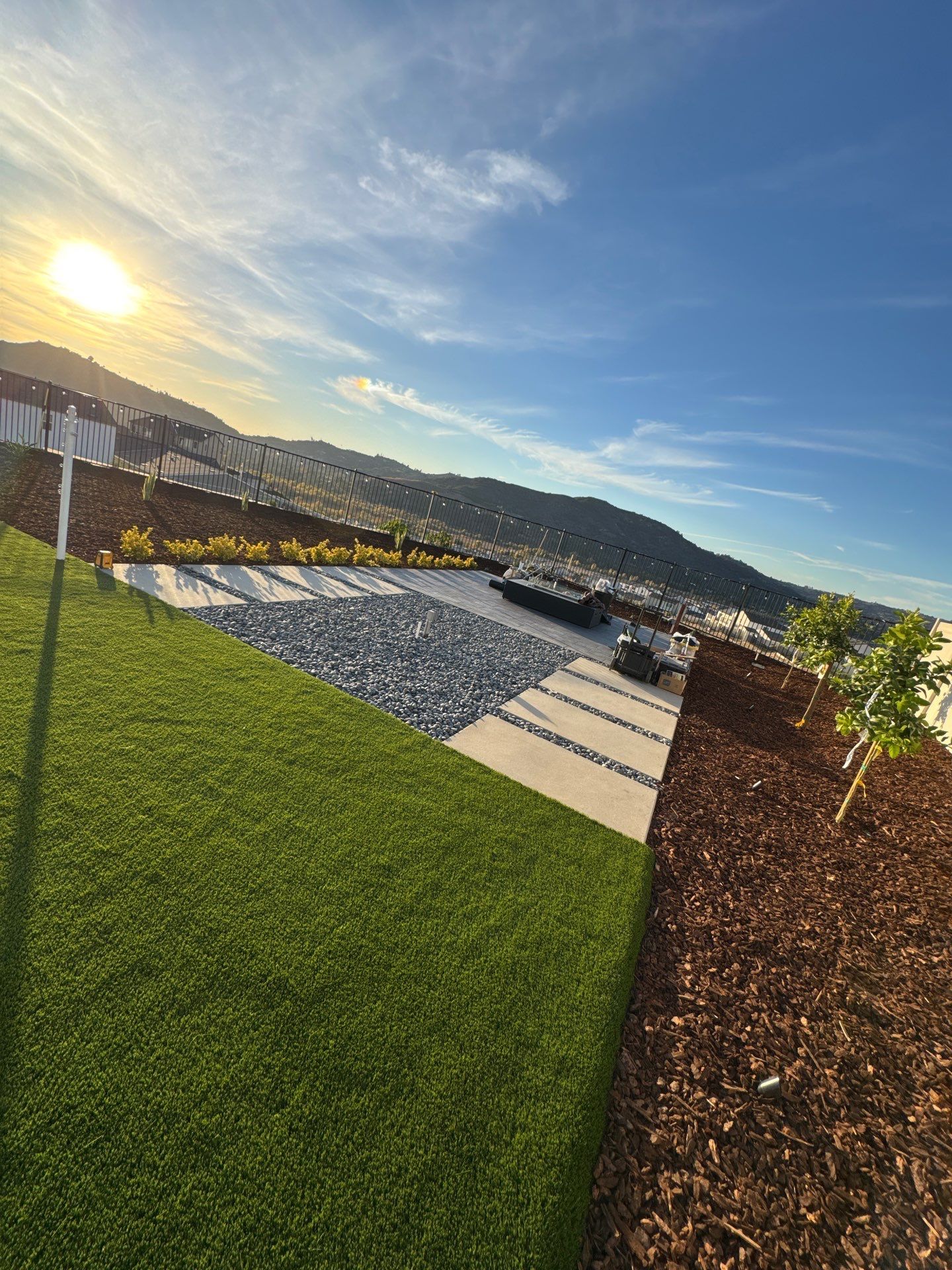 Green lawn, stone patio, and brown mulch border overlook a scenic hillside under a bright sun and blue sky.