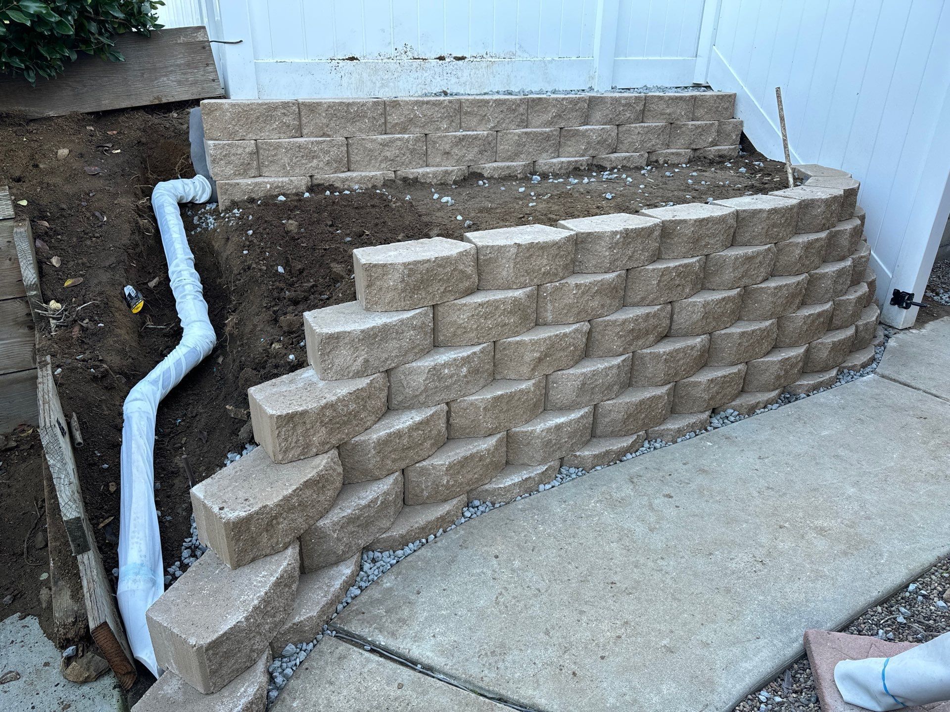 Retaining wall constructed with tan blocks, curved shape. White drainage pipe. Dirt, gravel, and concrete path visible.
