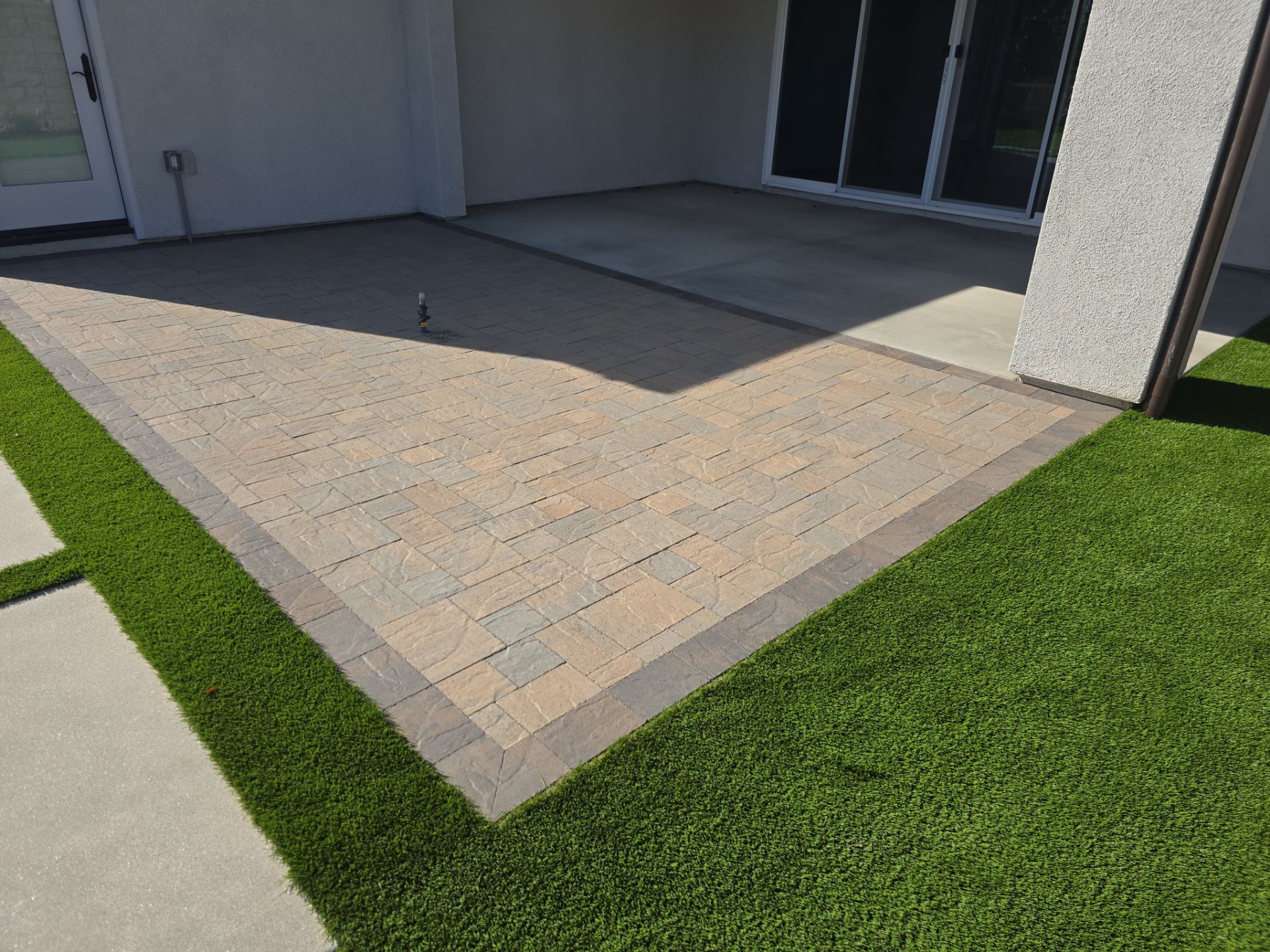 Brick patio with dark border, adjacent to green grass and light-colored building.