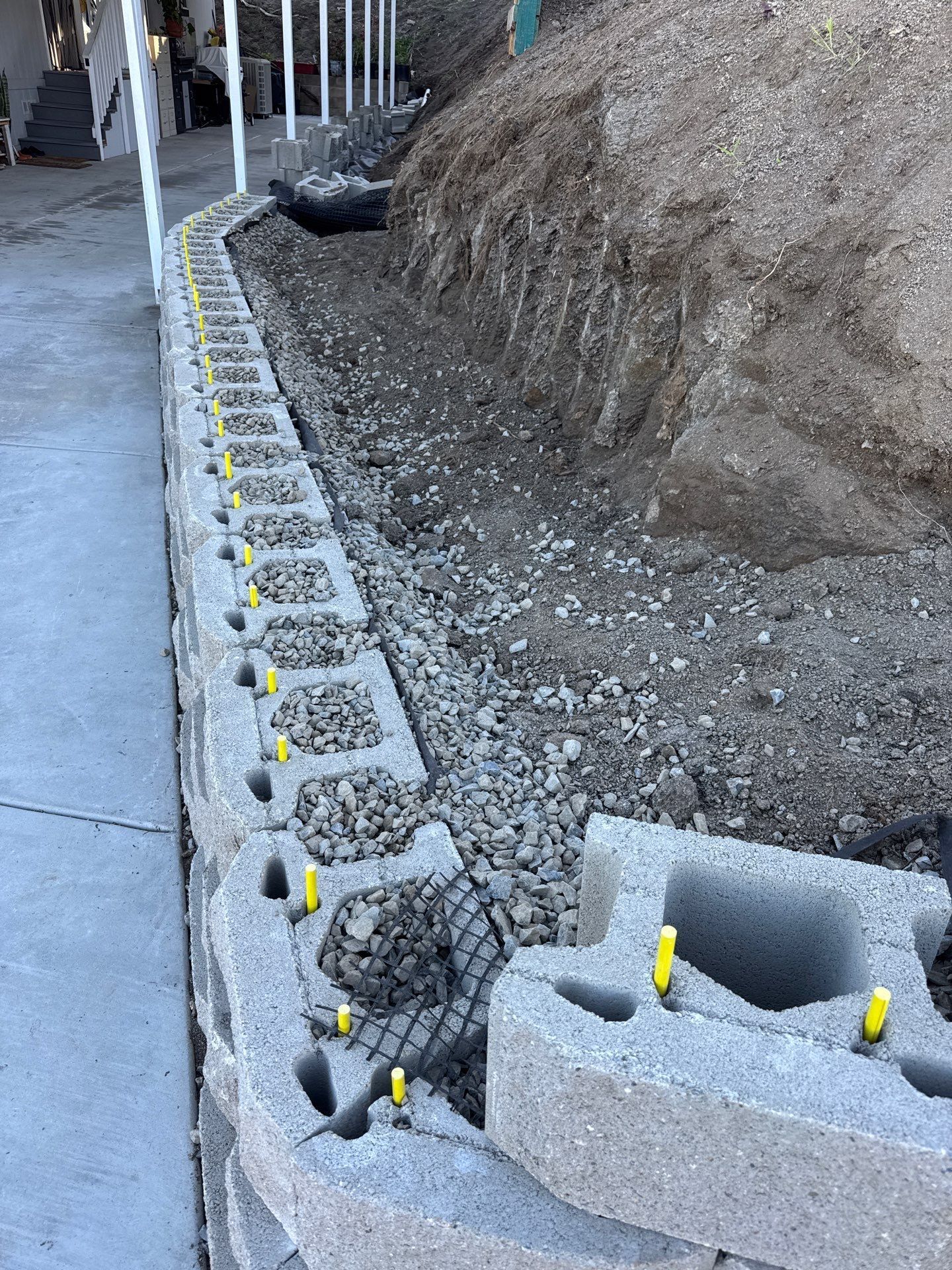 Retaining wall under construction with gray blocks, gravel, and dirt. Yellow pins hold blocks.