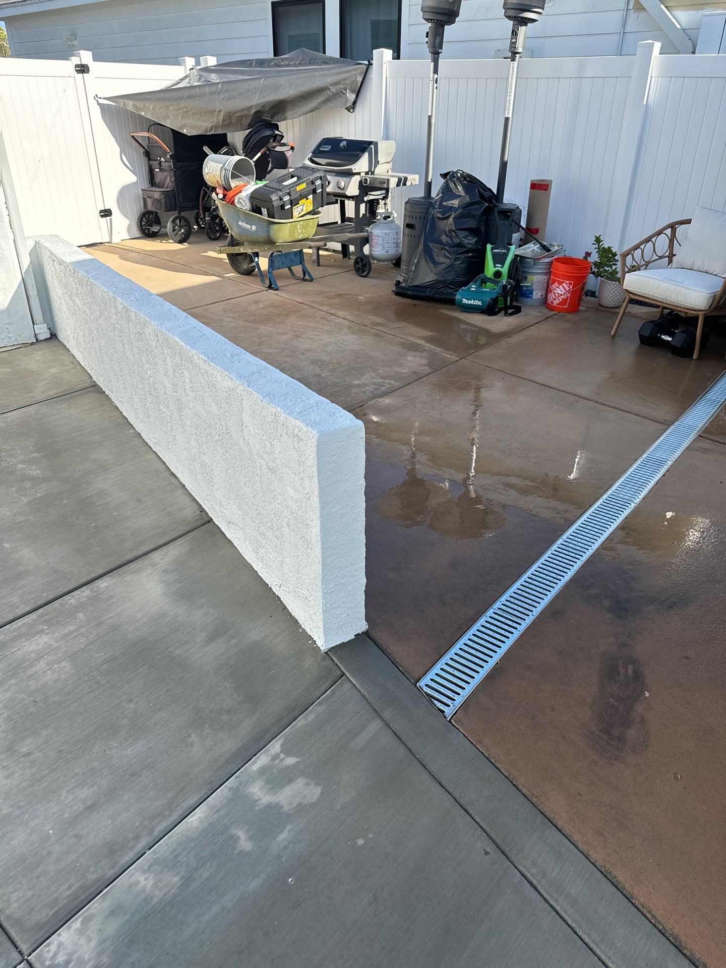 Concrete patio with textured wall, drainage channel. Tools, grill, and patio furniture visible.