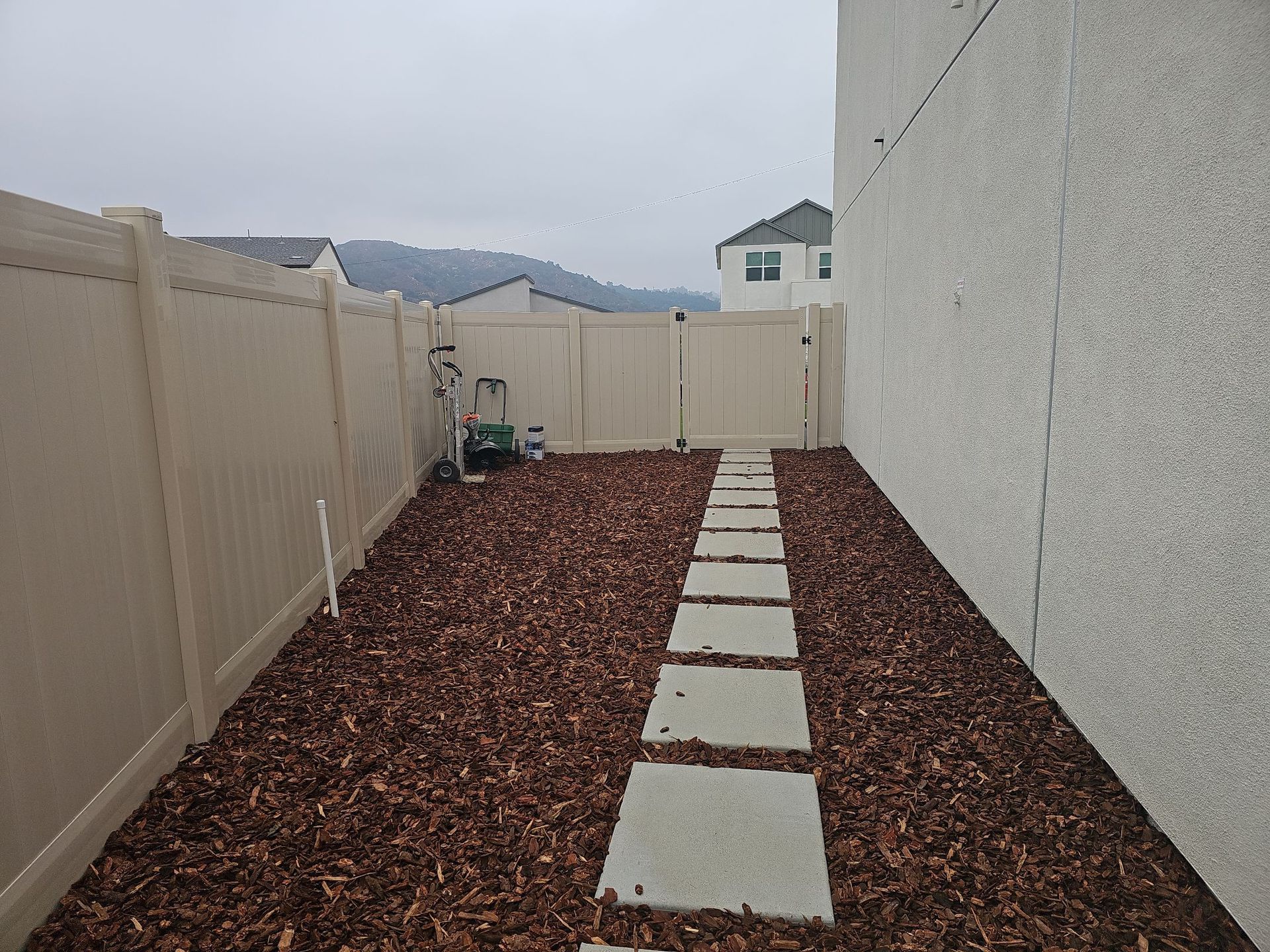 A narrow backyard with a stone path, beige fences, wood chips, and a building on the right.