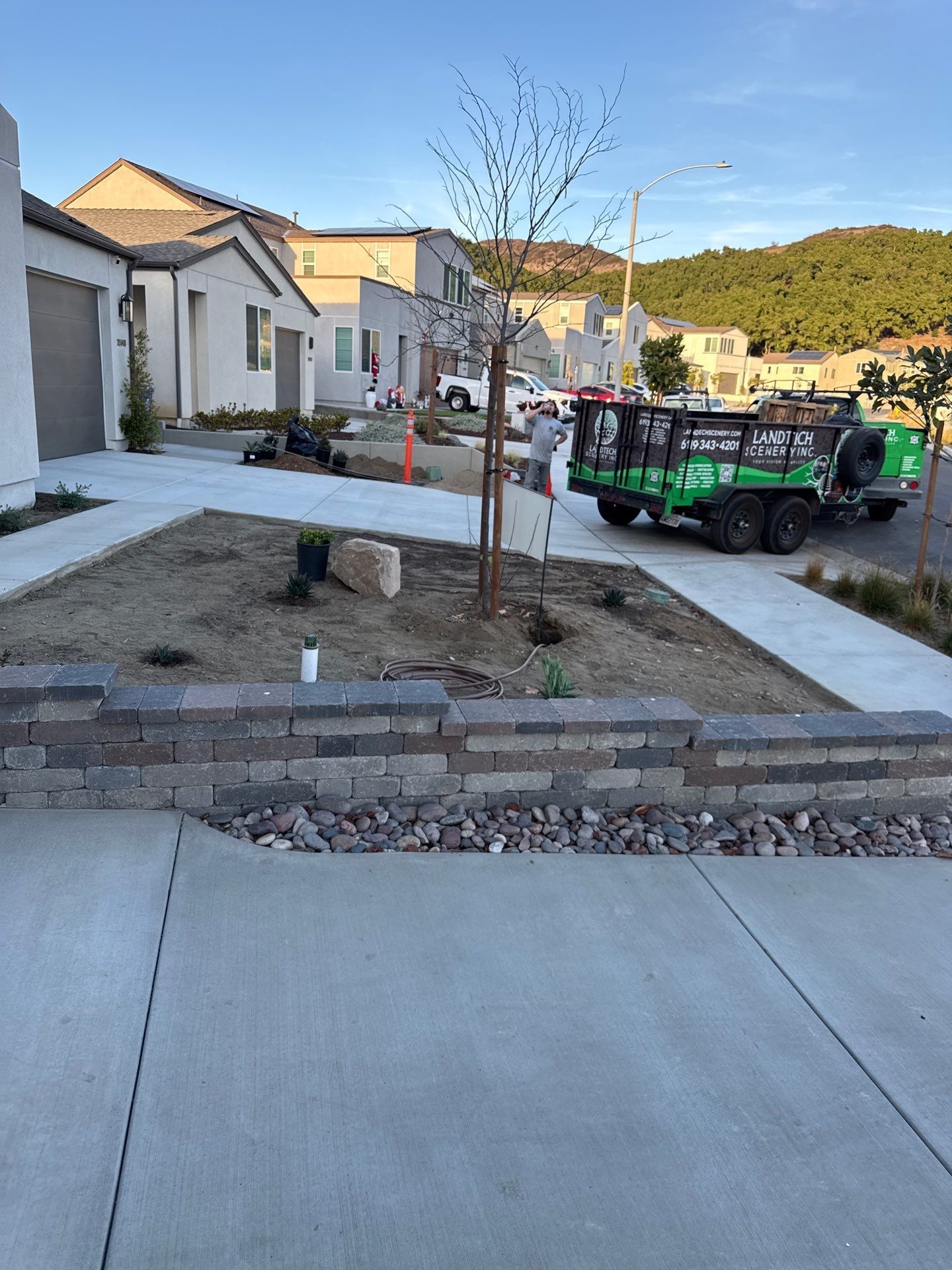 Residential landscaping in progress, featuring a retaining wall, bare tree, and green truck. Houses and hills in the background.