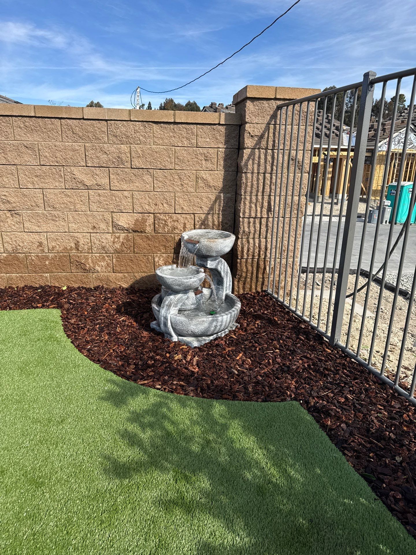 Water fountain in a landscaped corner with a brick wall, metal fence, and green turf.