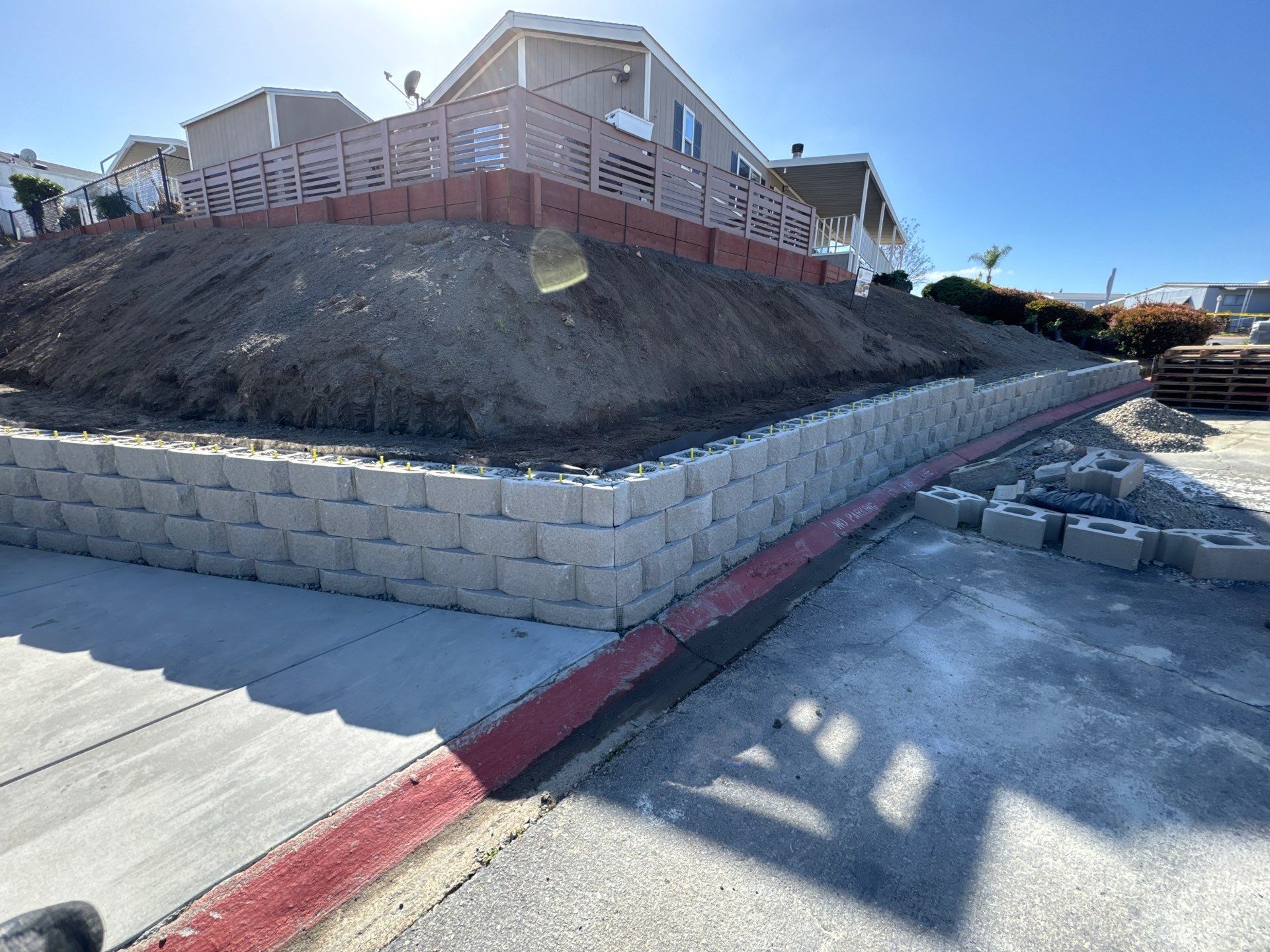 Retaining wall construction with gray blocks; a hill, building, and asphalt in the background. Sunny day.