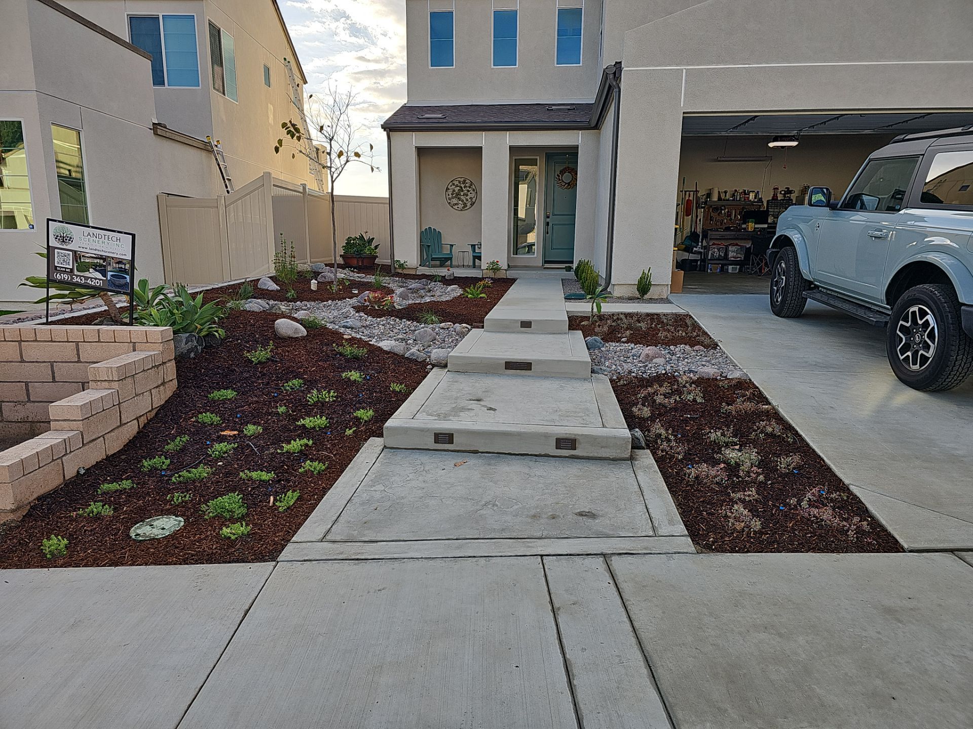 Two-story house with landscaped front yard; concrete pathway leads to the entrance. A blue truck is parked in the garage.