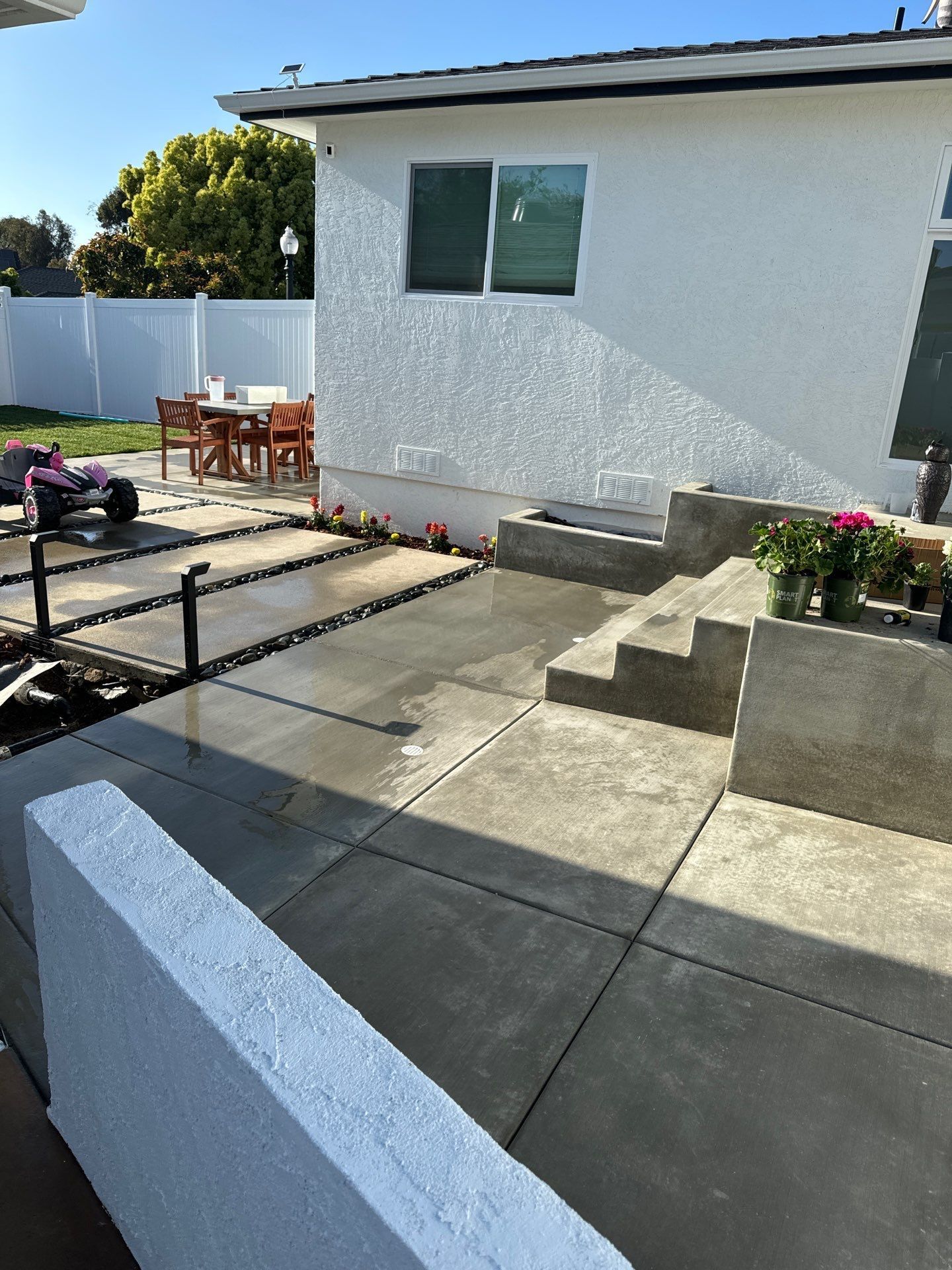 Exterior view of a patio with concrete flooring, steps, and a white stucco wall. Wooden furniture sits in the background.