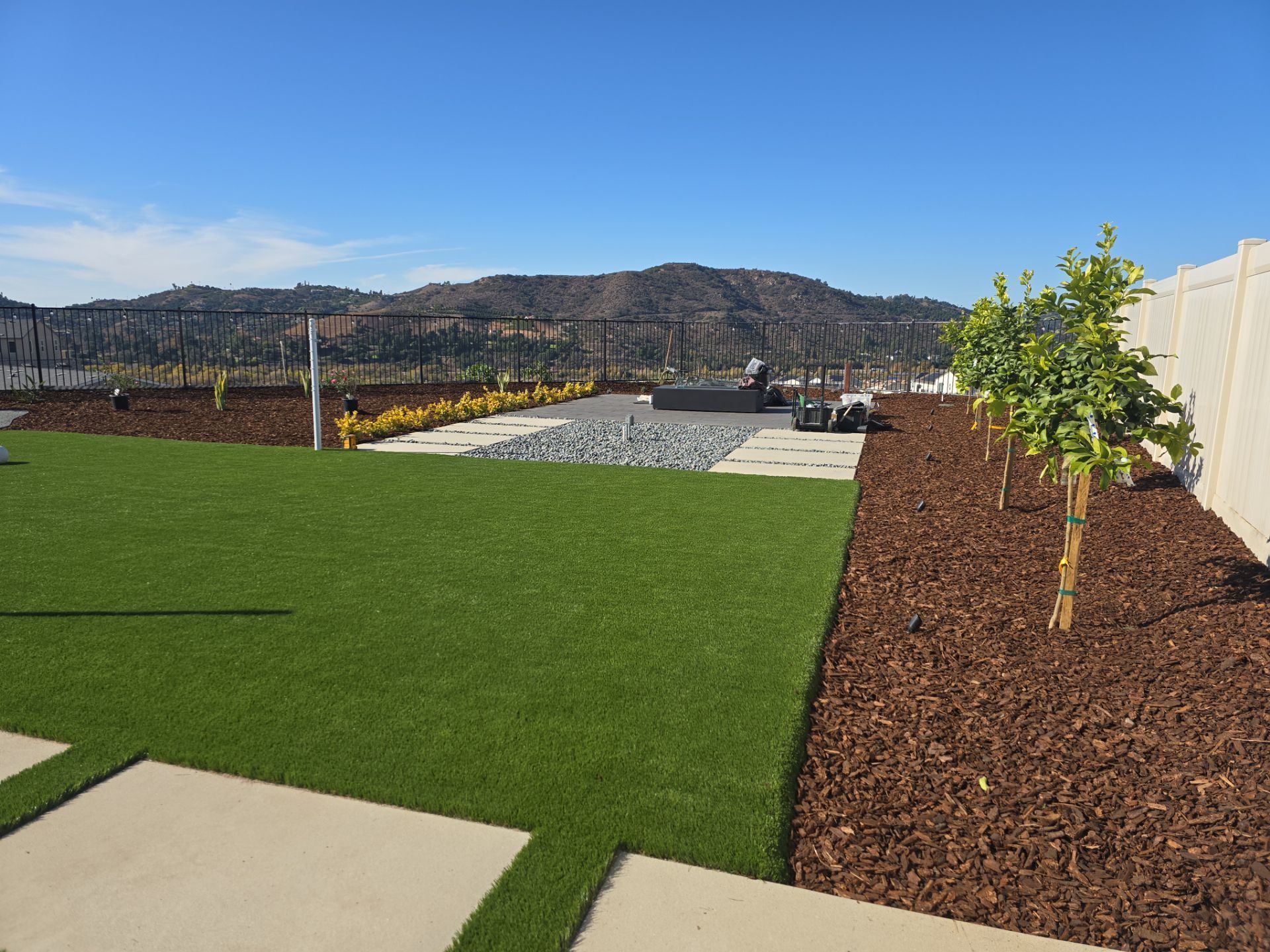 Green artificial lawn with brown mulch borders, trees, and mountain backdrop under a blue sky.
