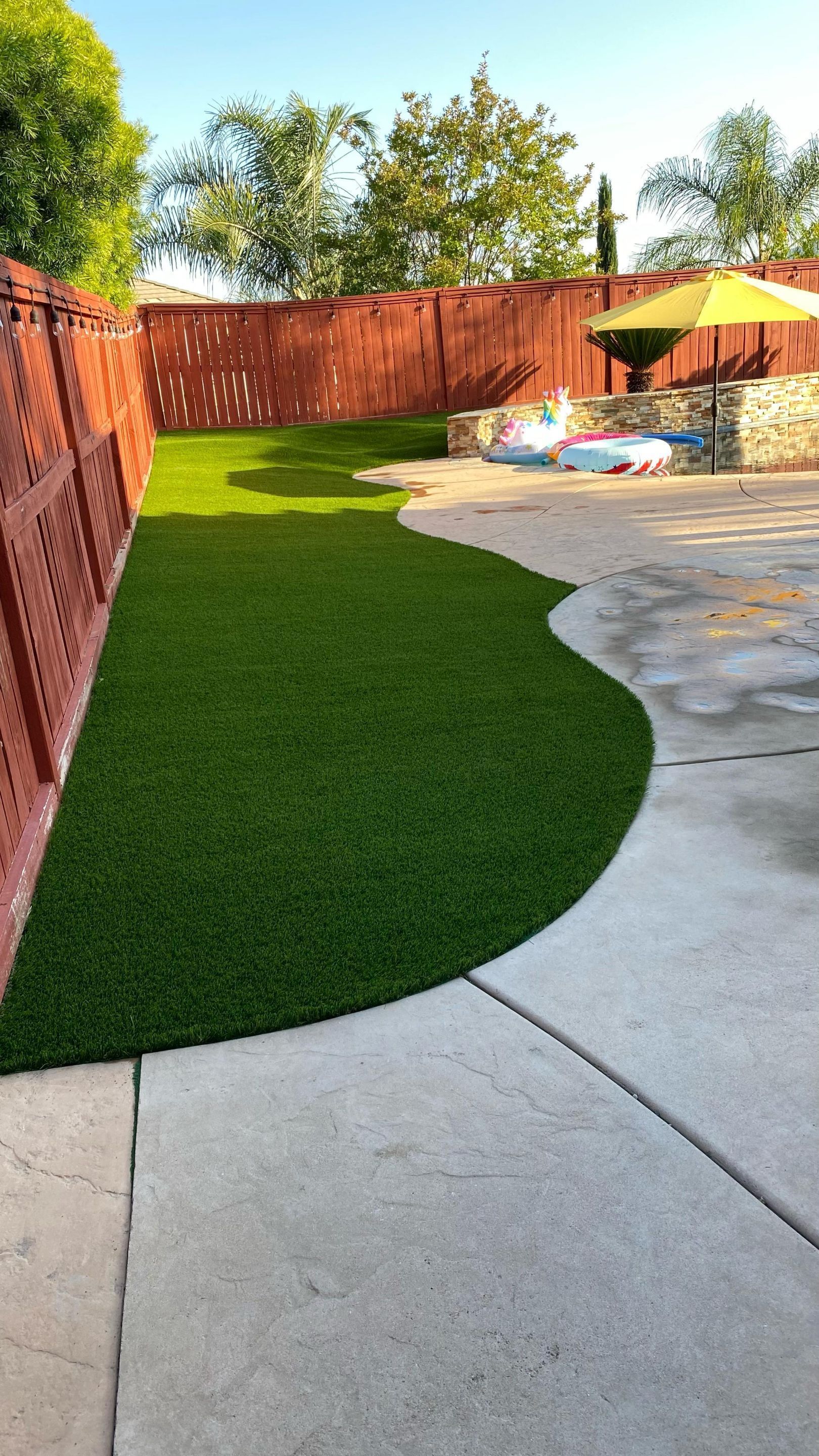 Green artificial grass in a backyard with a red fence, concrete patio, and umbrella.