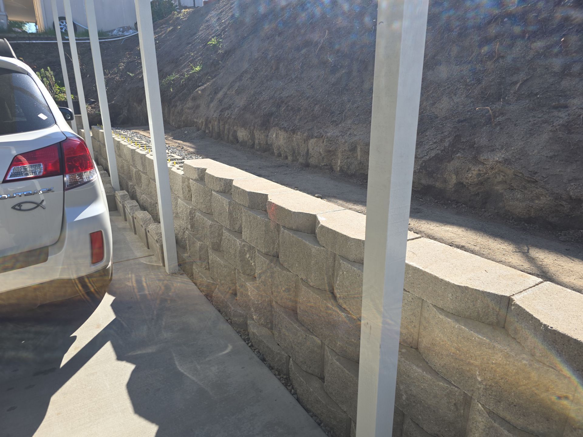 White car parked next to a retaining wall made of stacked concrete blocks under a white carport.