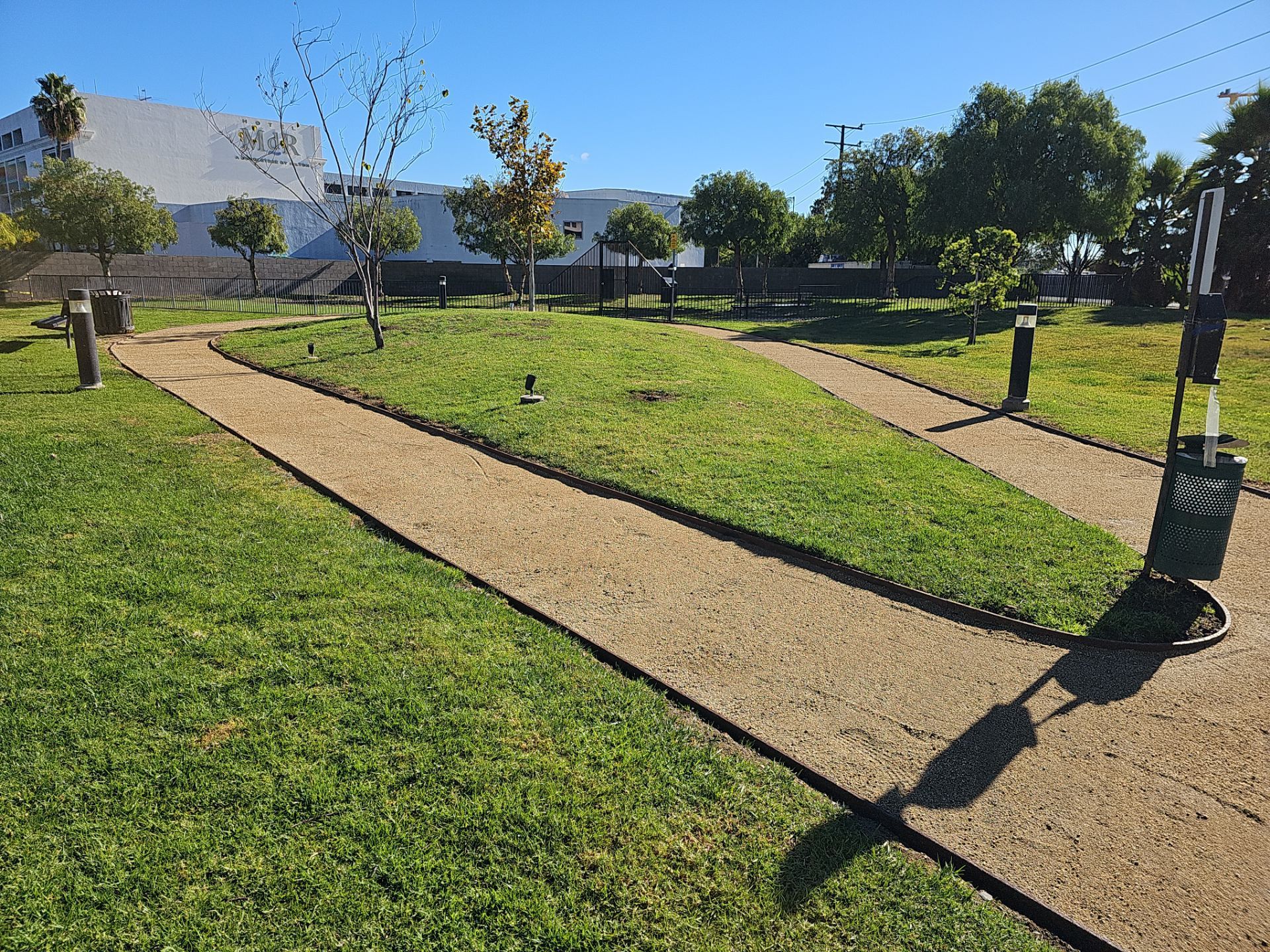 Gravel paths through a grassy park under a blue sky, with trees, lamps, and a building in the background.