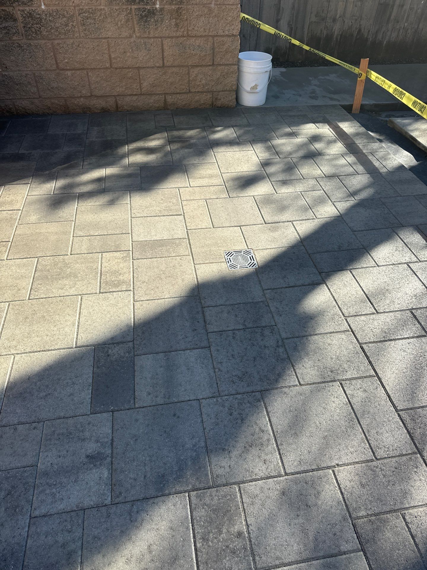 Paver patio with a building wall, bucket, caution tape, and a long shadow.