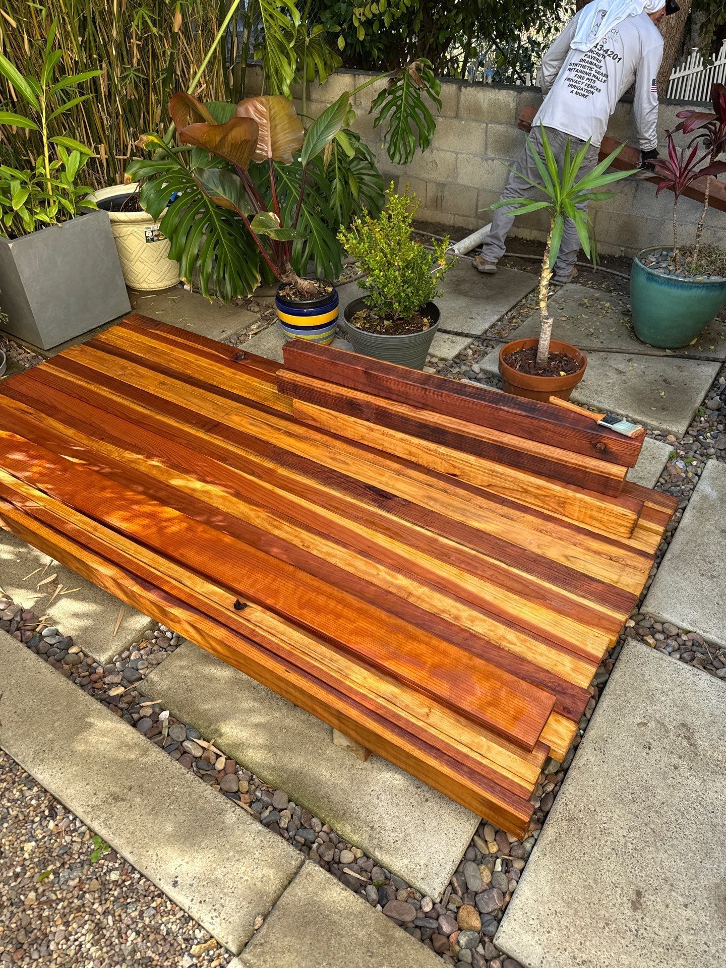 Wooden planks laid out on a stone patio with a person working in the background amidst potted plants.