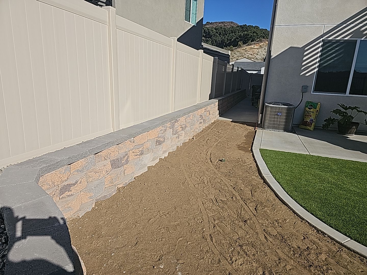 Retaining wall with beige pavers and tan ground next to a beige fence, pathway, and green lawn.