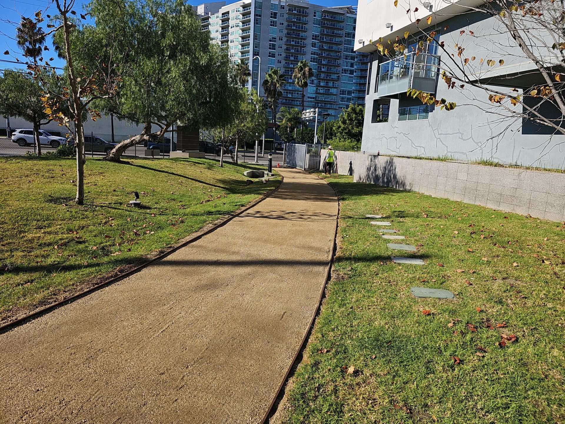 Gravel path through a grassy park with trees, leading toward modern buildings under a sunny sky.
