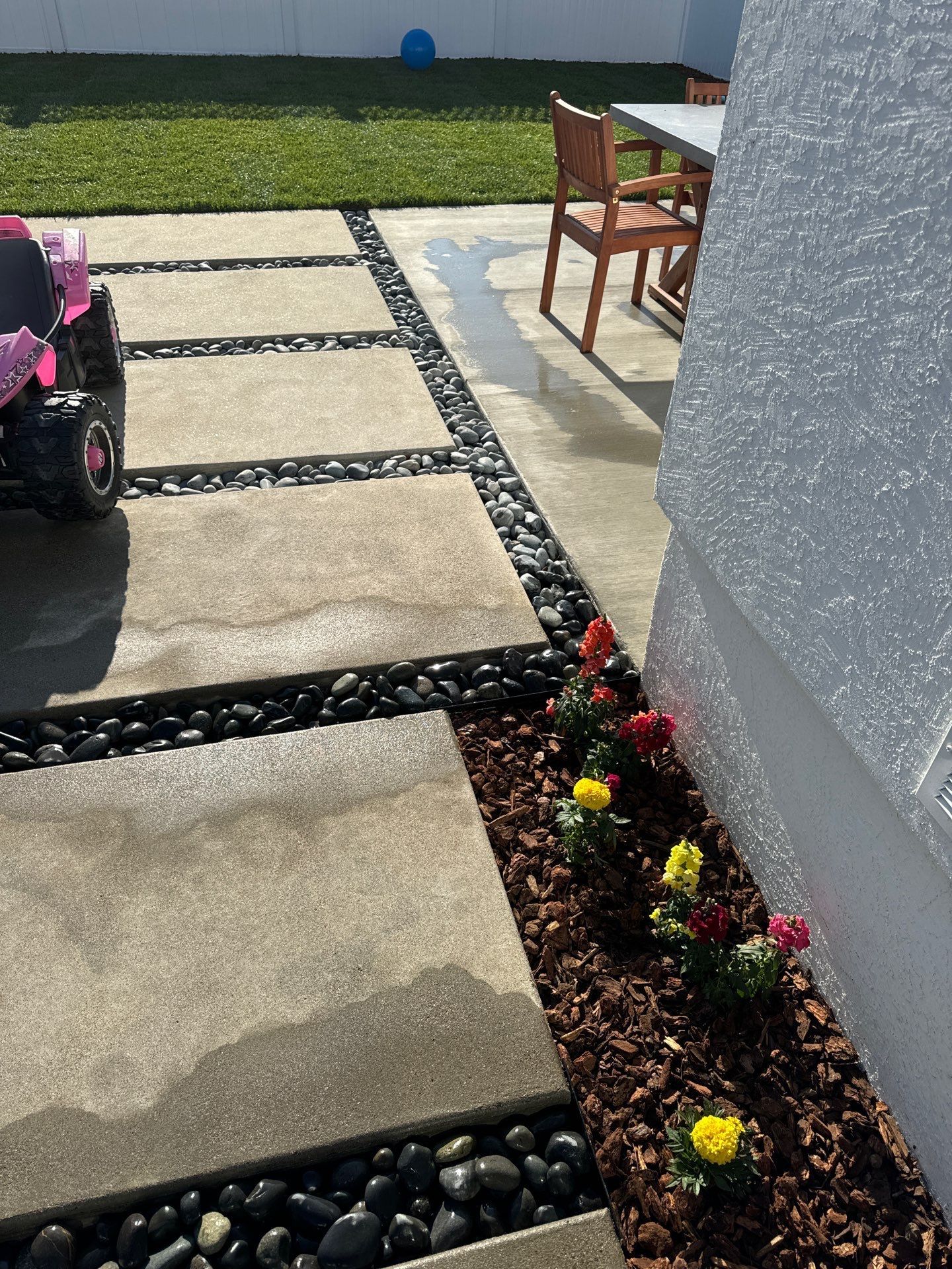 Square concrete pavers with black river rocks and flower bed. A child's toy is on the left.