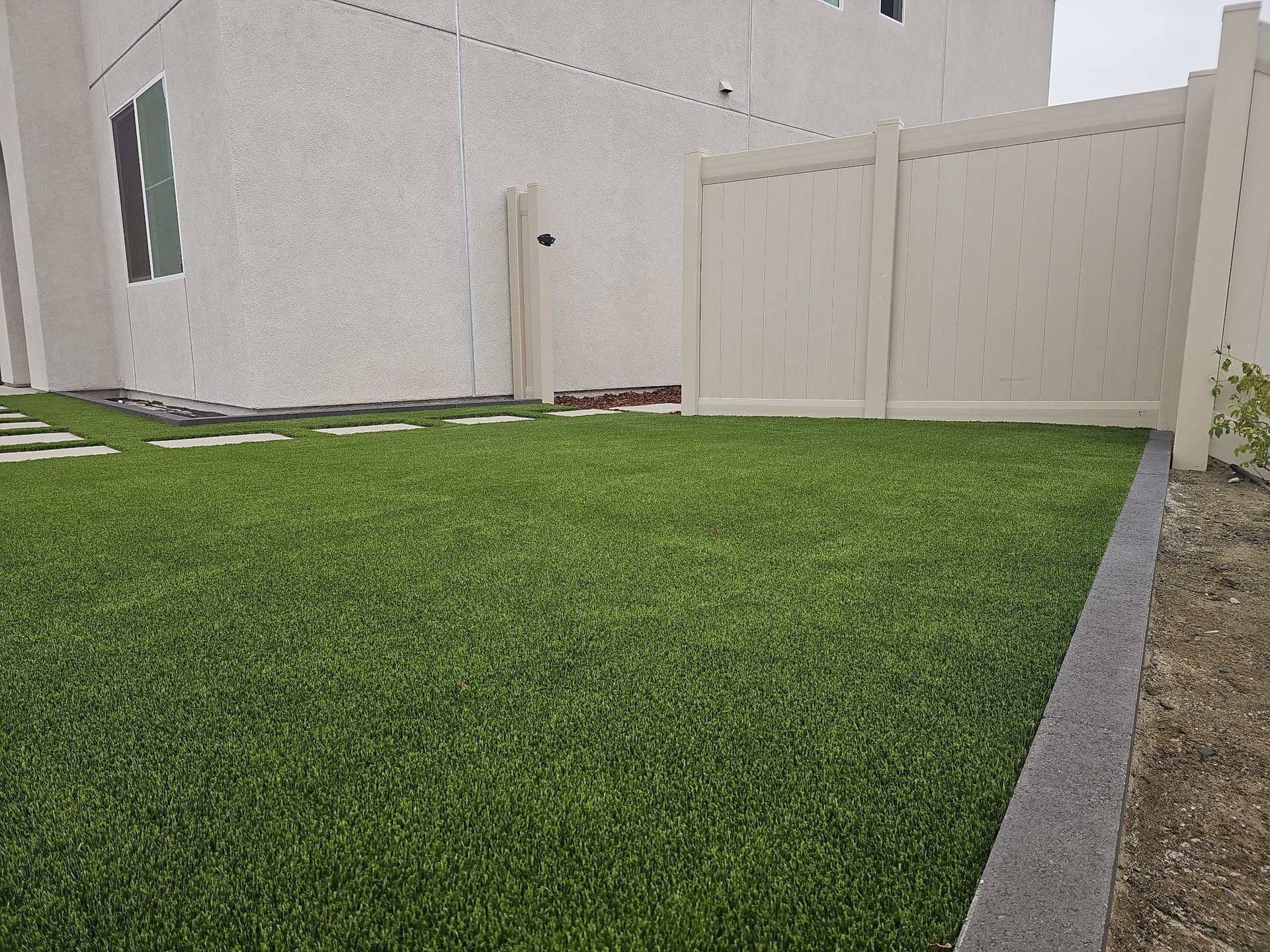 Artificial green grass in a backyard with a beige fence and a textured wall.