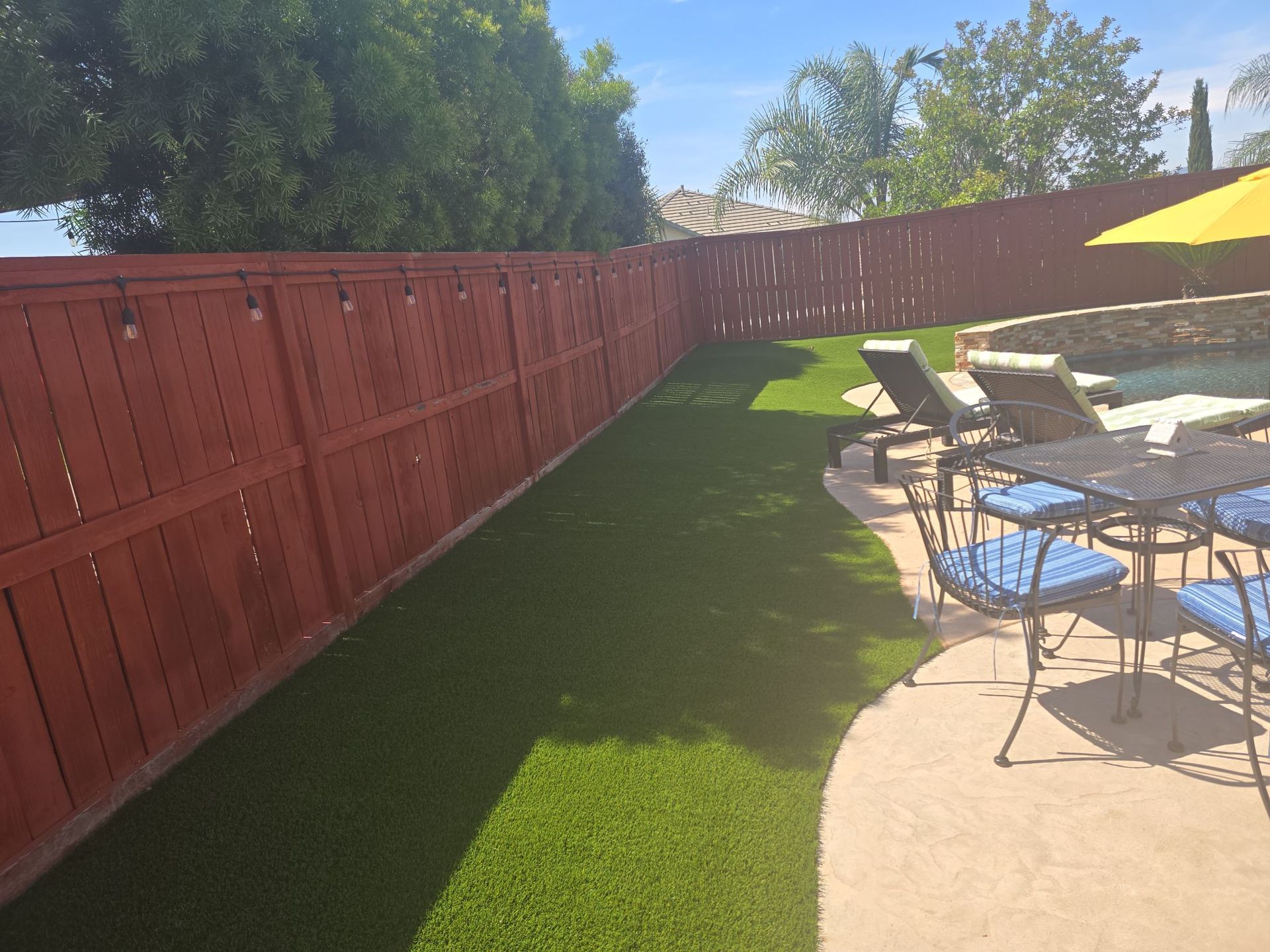 Red stained wooden fence alongside green artificial turf lawn, with pool and patio furniture visible.